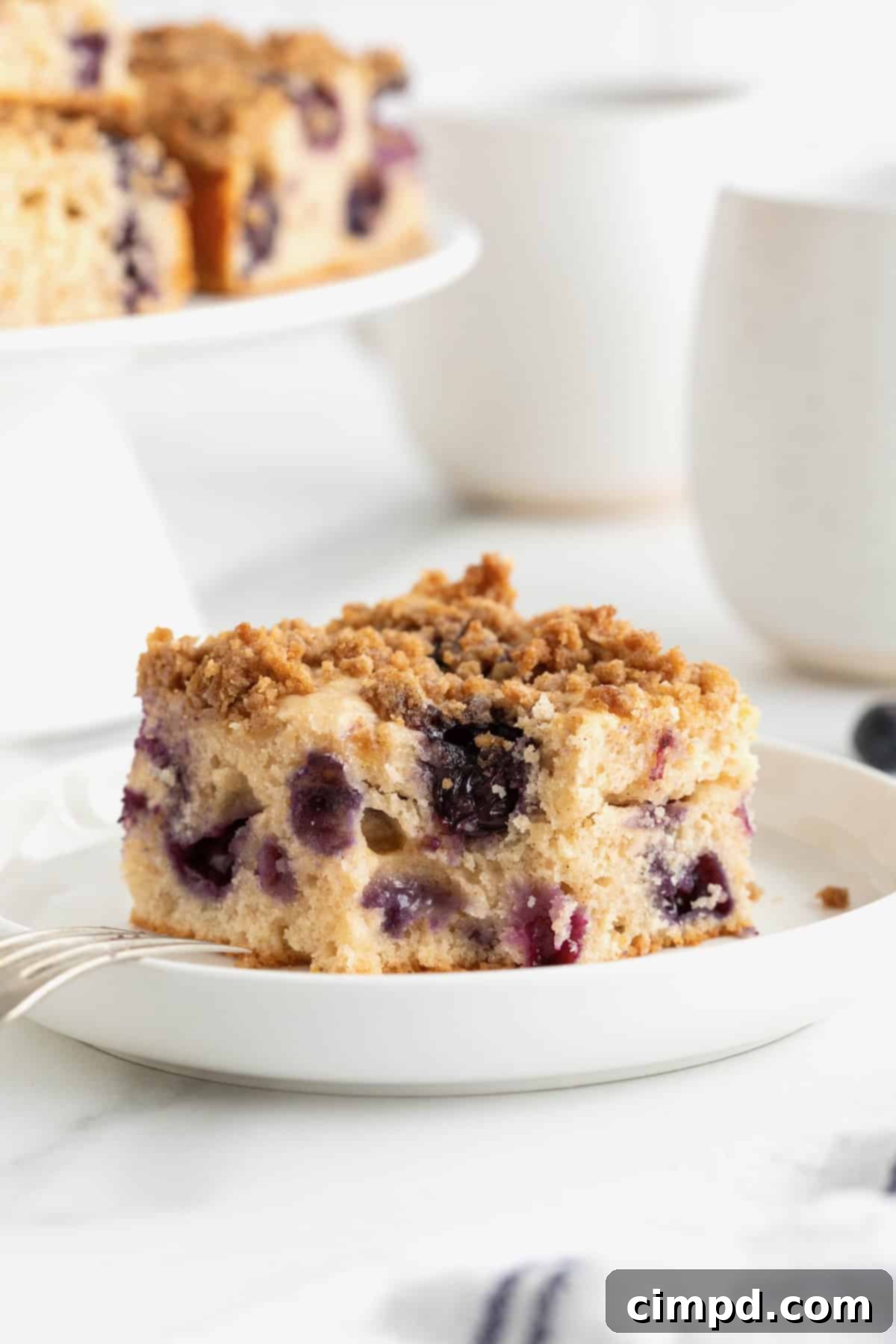 Blueberry Burst Greek Yogurt Crumb Cake 4 A piece of blueberry coffee cake on a white rimmed plate with a gold toned fork to the left.
