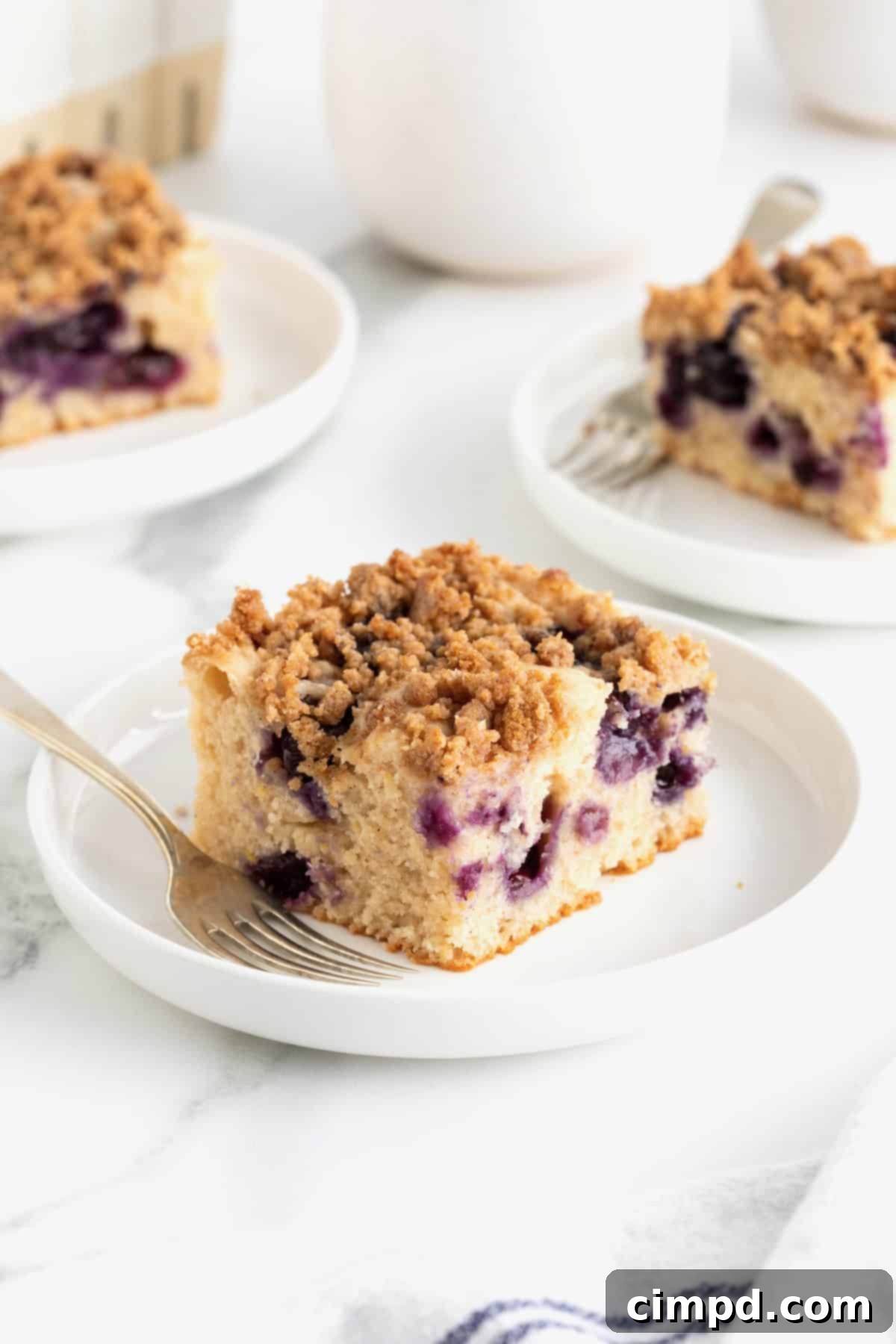 Blueberry Burst Greek Yogurt Crumb Cake 5 Three pieces of blueberry coffee cake on a white rimmed plates with a gold toned fork to the left arranged on a white marble counter.