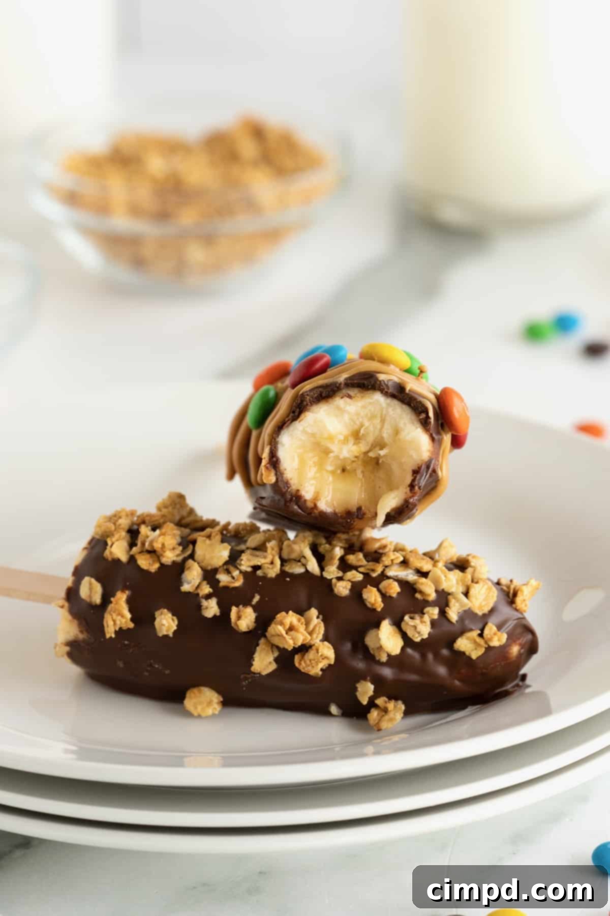 A chocolate covered frozen banana covered with M&Ms laying on a chocolate covered frozen banana coated in granola.