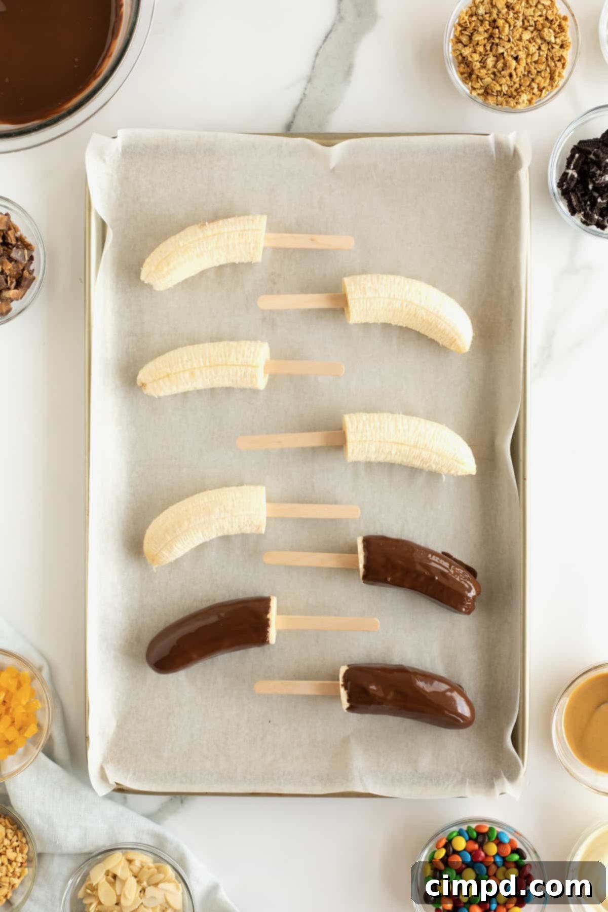 Bananas on popsicle sticks covered in melted chocolate on a parchment lined baking tray.