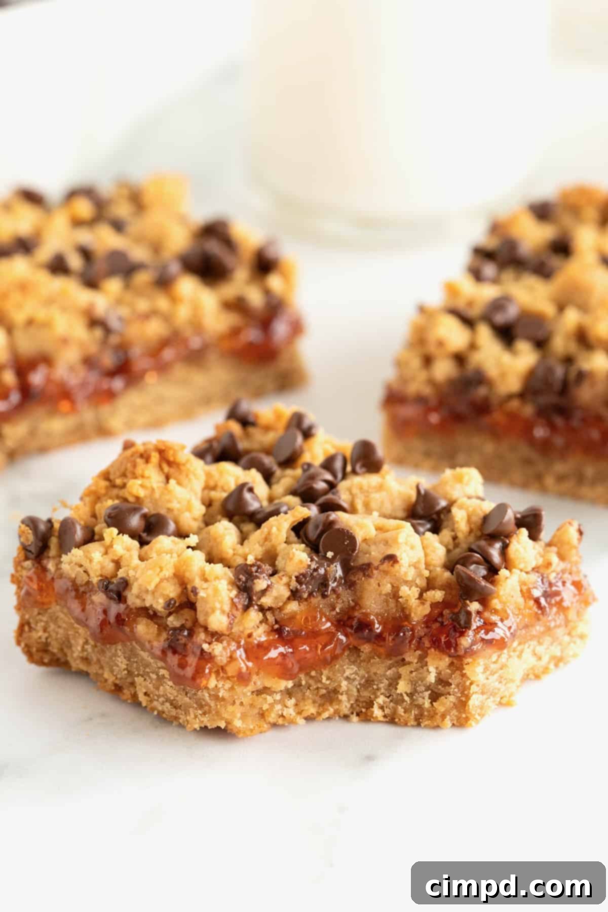 Three peanut butter and jelly bars topped with mini chocolate chips on a white surface. The front bar has a bite out of it.