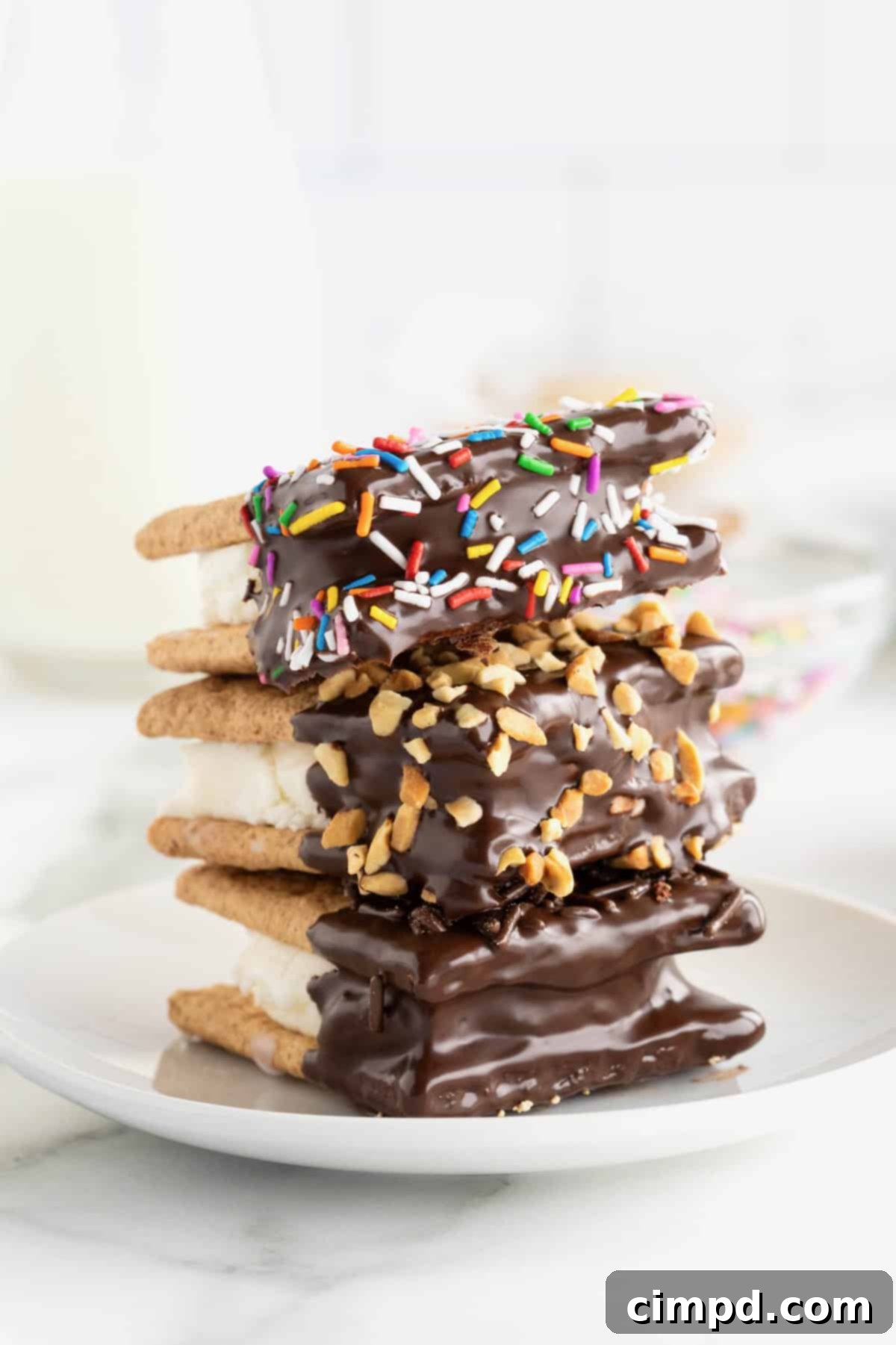 A stack of three chocolate dipped graham cracker ice cream sandwiches with sprinkles and nuts on a small white plate.