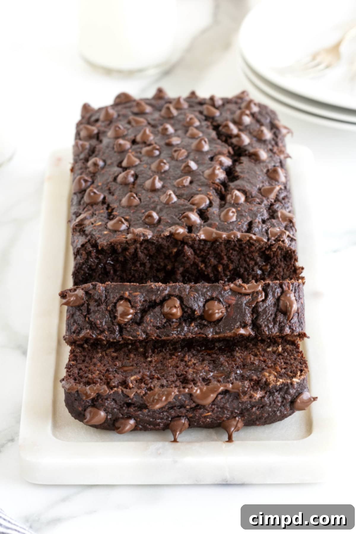 A freshly baked loaf of Flourless Chocolate Banana Bread, generously studded with melted chocolate chips on top. It sits elegantly on a white cutting board, set against a pristine white marble counter, inviting a delicious slice.