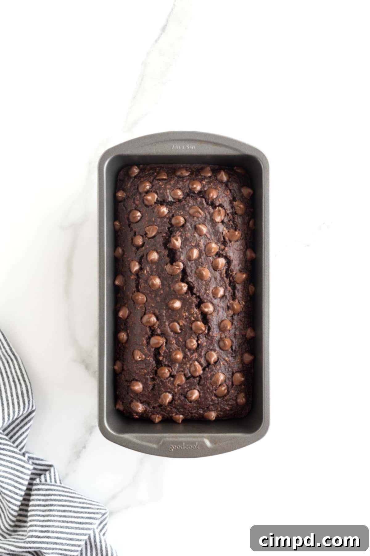 A perfectly baked loaf of Flourless Chocolate Banana Bread, beautifully golden and studded with glistening chocolate chips, rests in a dark metal loaf pan on a cool white marble counter.