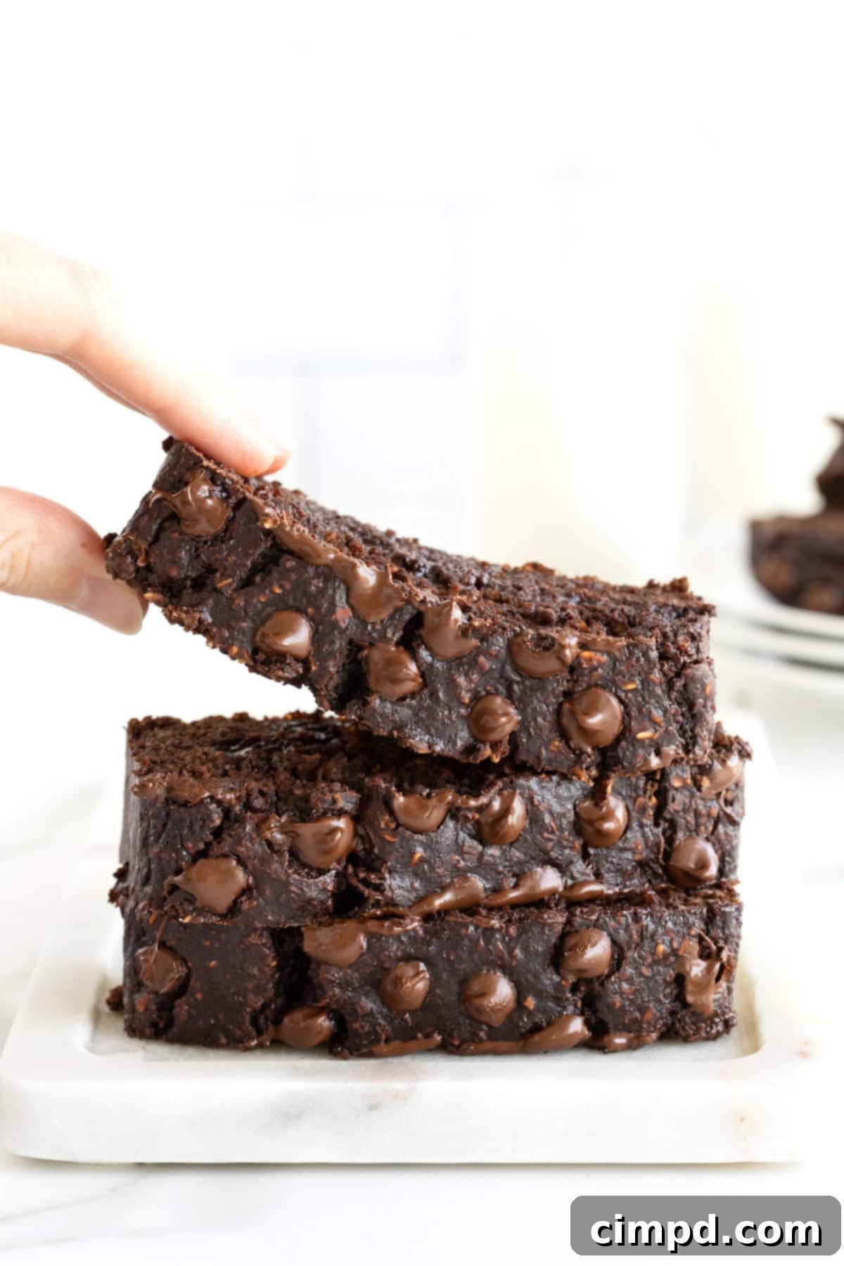 A hand gently lifts a warm, thick slice of chocolate banana bread from a stack of three inviting slices, arranged beautifully on a small white plate.