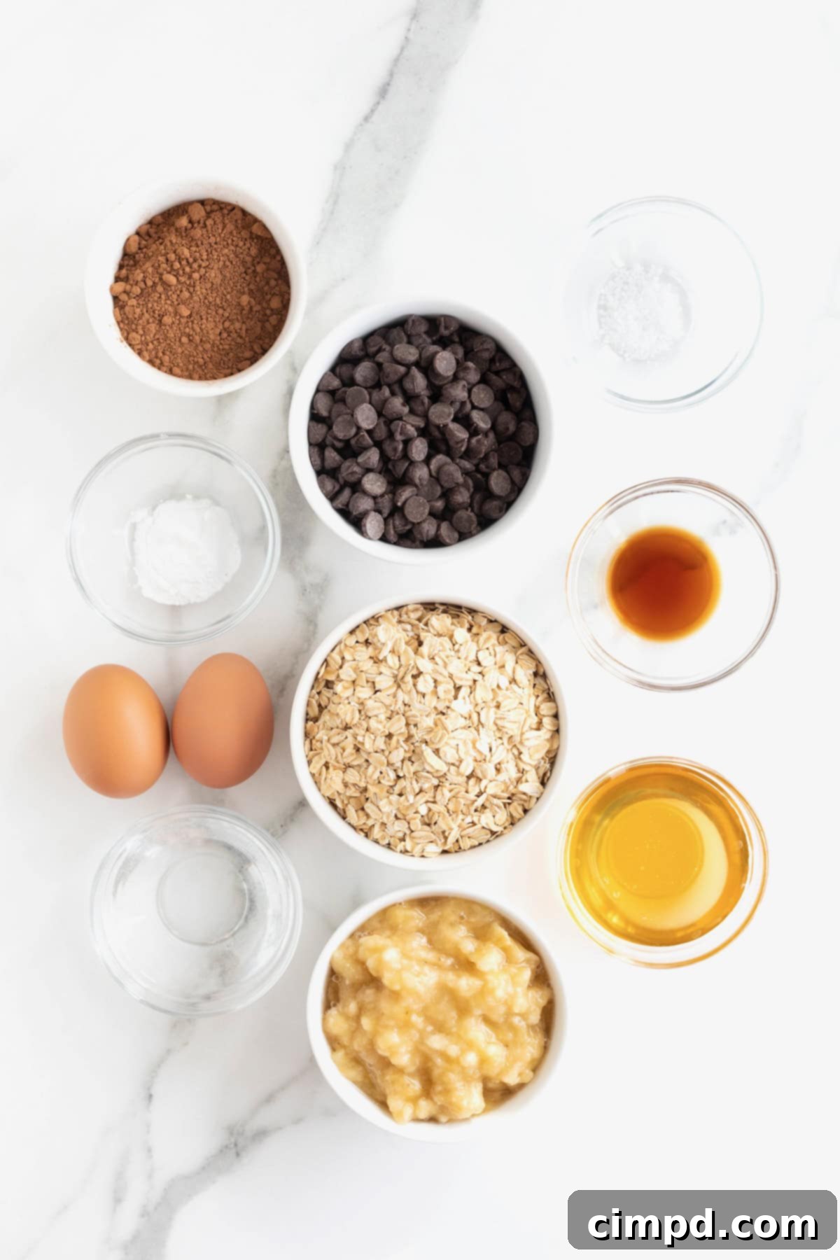 A beautifully arranged collection of fresh ingredients ready to make Flourless Chocolate Banana Bread, displayed in small white glass dishes on a pristine white marble counter, highlighting their natural appeal.