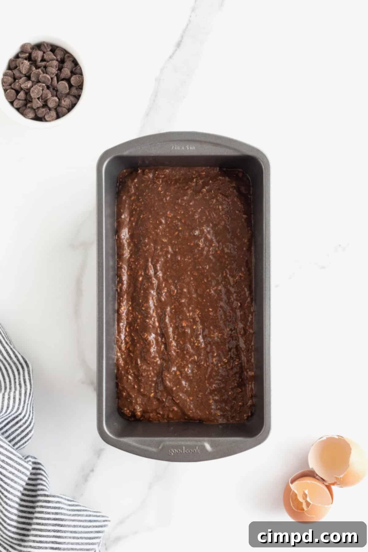Thick, rich chocolate banana bread batter fills a dark aluminum loaf pan, perfectly prepared and ready for baking, resting on a clean white marble counter.