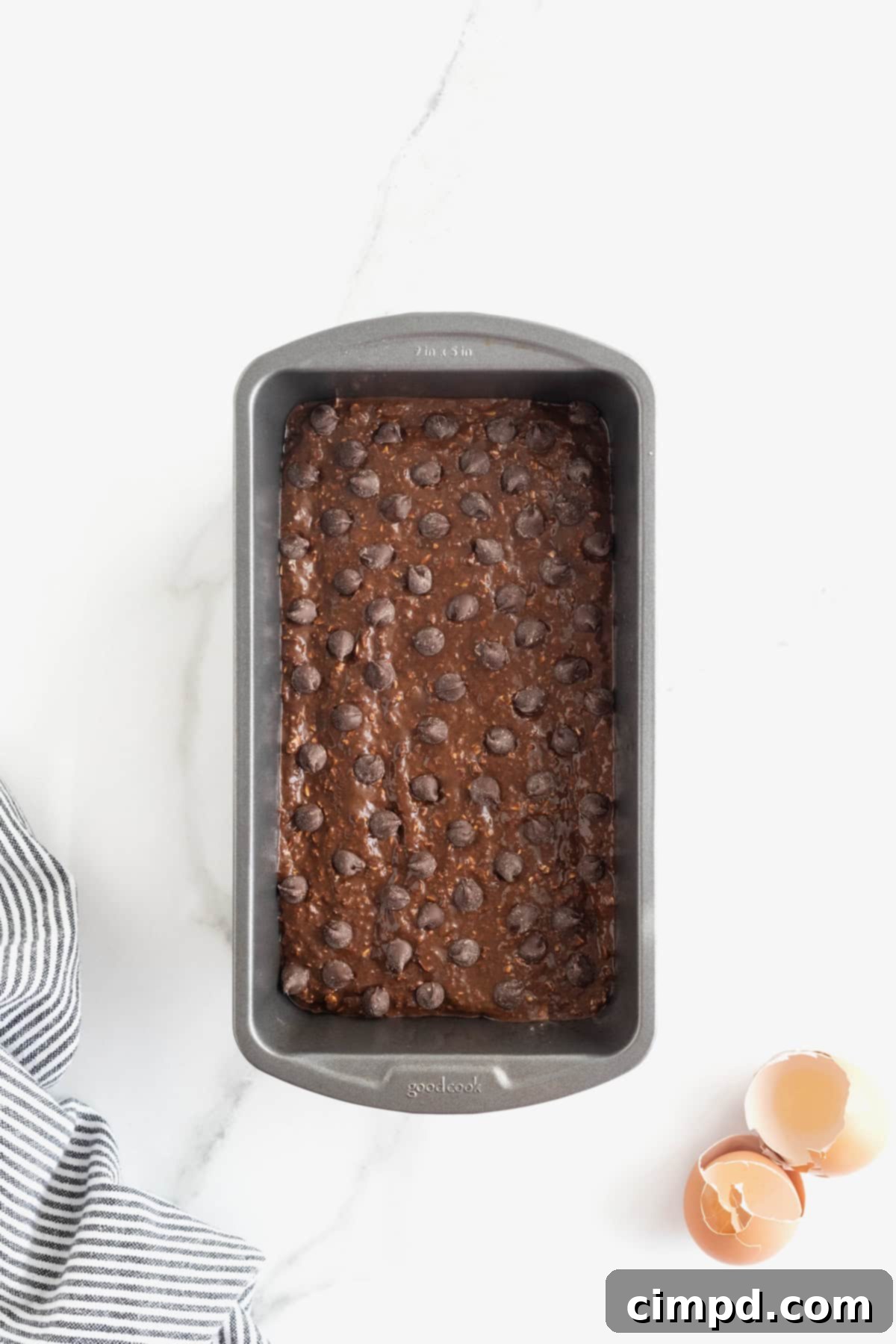 Dark, luscious chocolate banana bread batter, generously topped with an extra sprinkle of chocolate chips, sits in a dark aluminum loaf pan, eagerly awaiting its transformation in the oven.