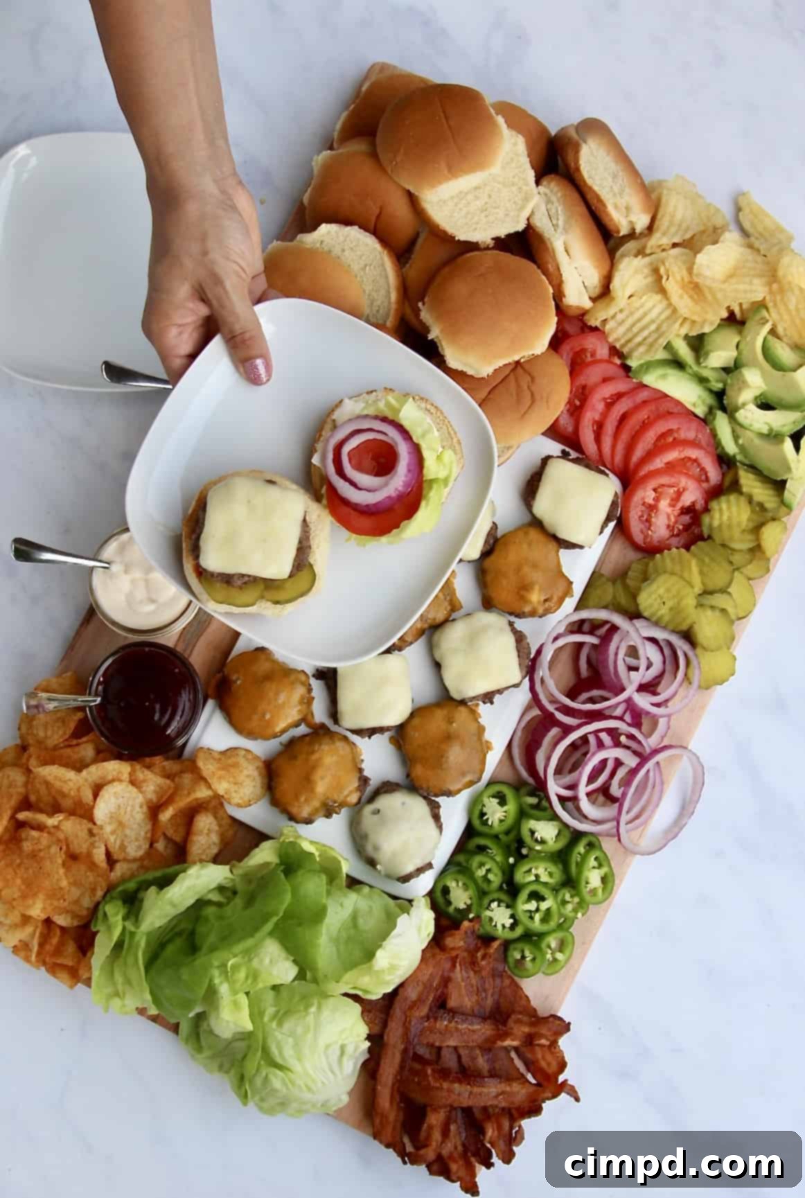 A top-down view of a Build-Your-Own Burger Board filled with fresh ingredients like avocado, tomatoes, various cheeses, and different sauces in small jars.