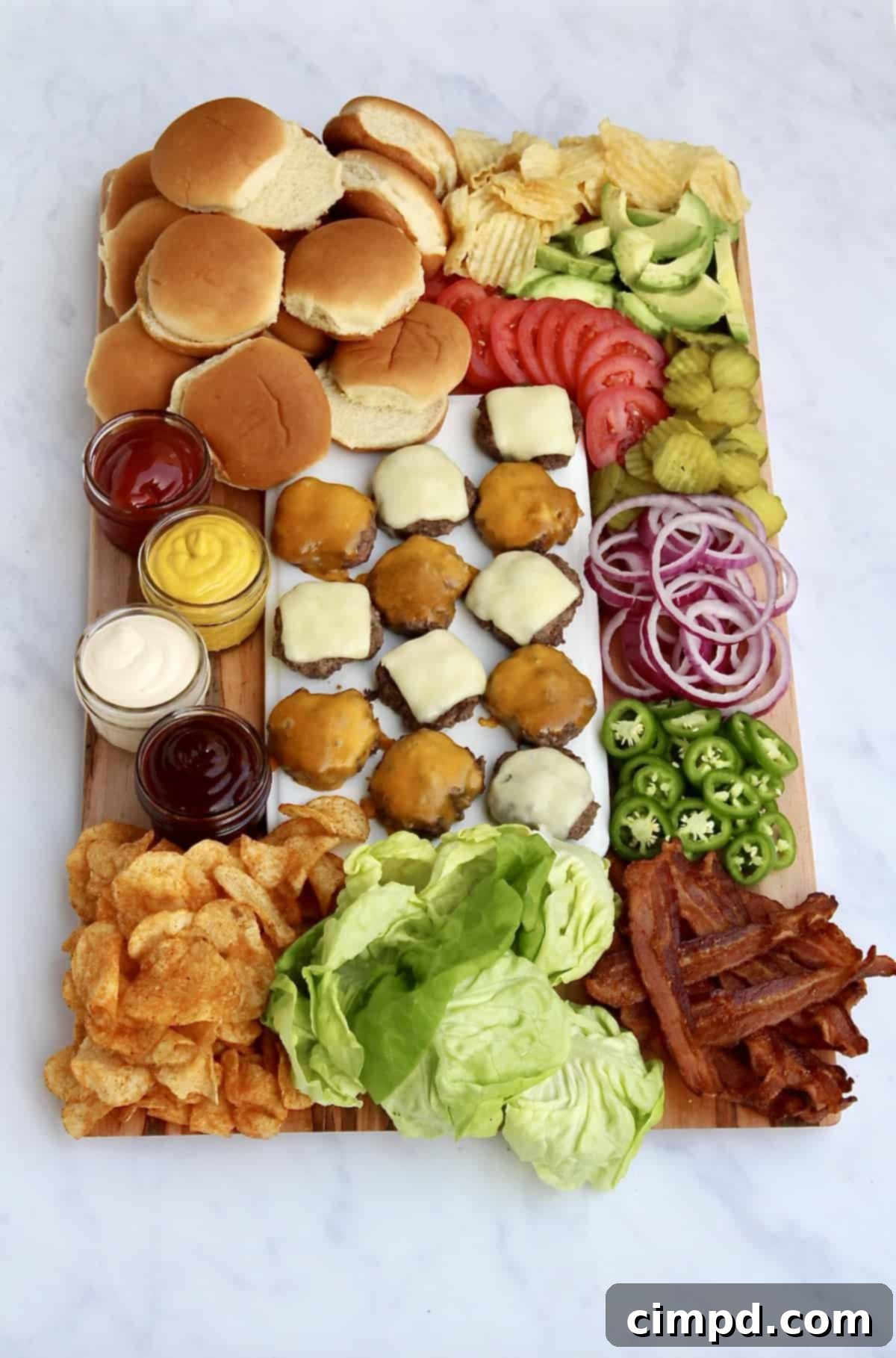 A fully assembled Build-Your-Own Burger Board by The BakerMama, showing grilled burger patties in the center surrounded by an abundance of fresh, colorful toppings.