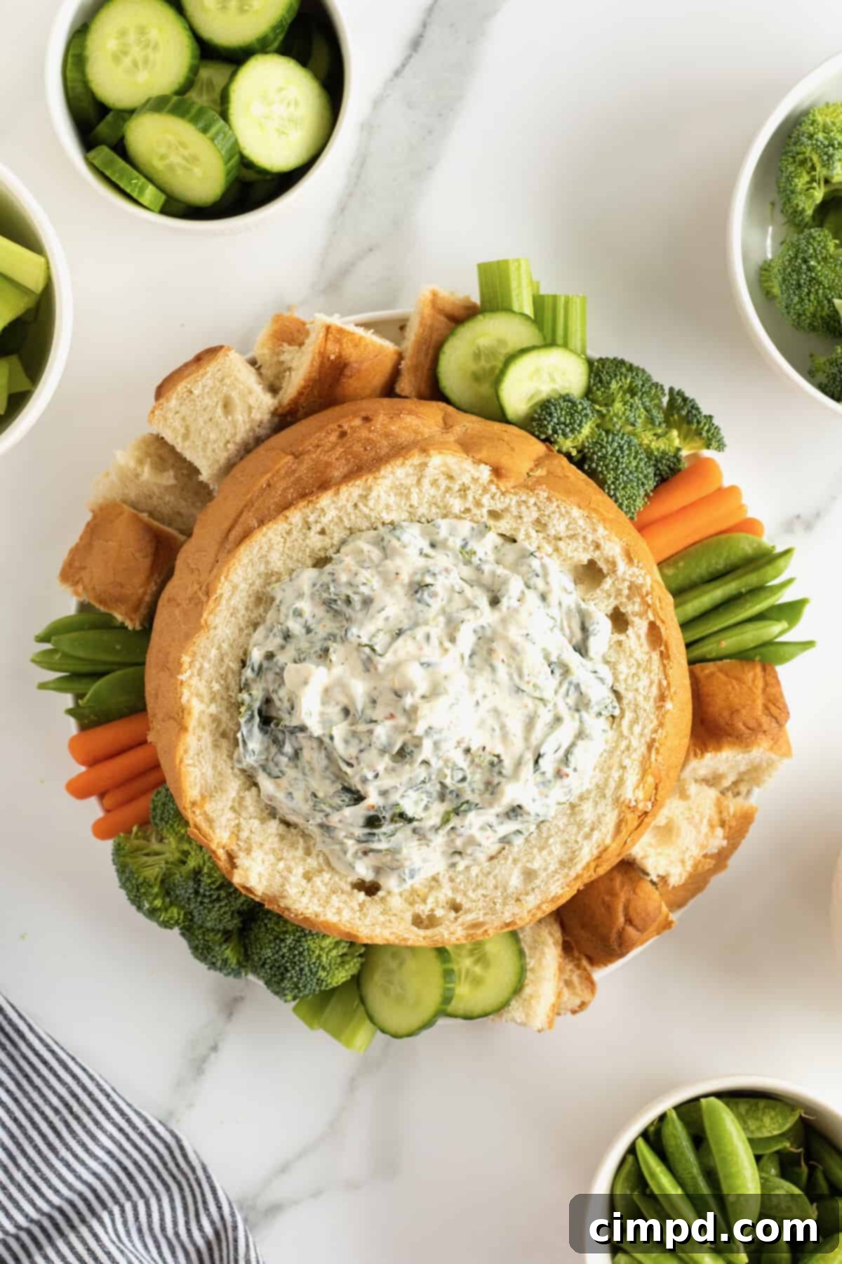 Creamy spinach dip in a bread bowl surrounded by fresh vegetables and bread chunks. A visually appealing appetizer for any occasion.
