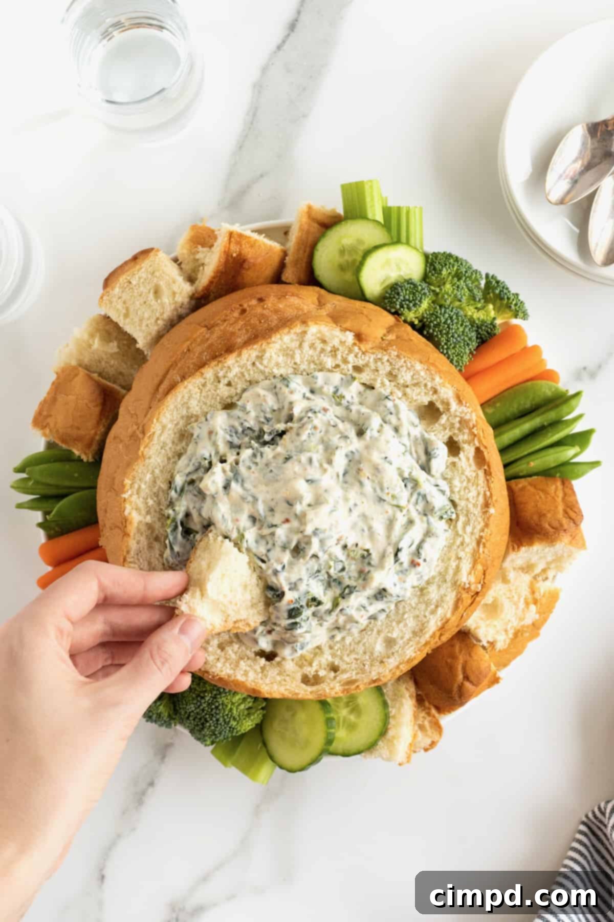Creamy spinach dip presented in a bread bowl, surrounded by an appetizing display of fresh vegetables and bread chunks.