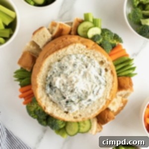 Creamy spinach dip in bread bowl surrounded by fresh vegetables and bread chunks.