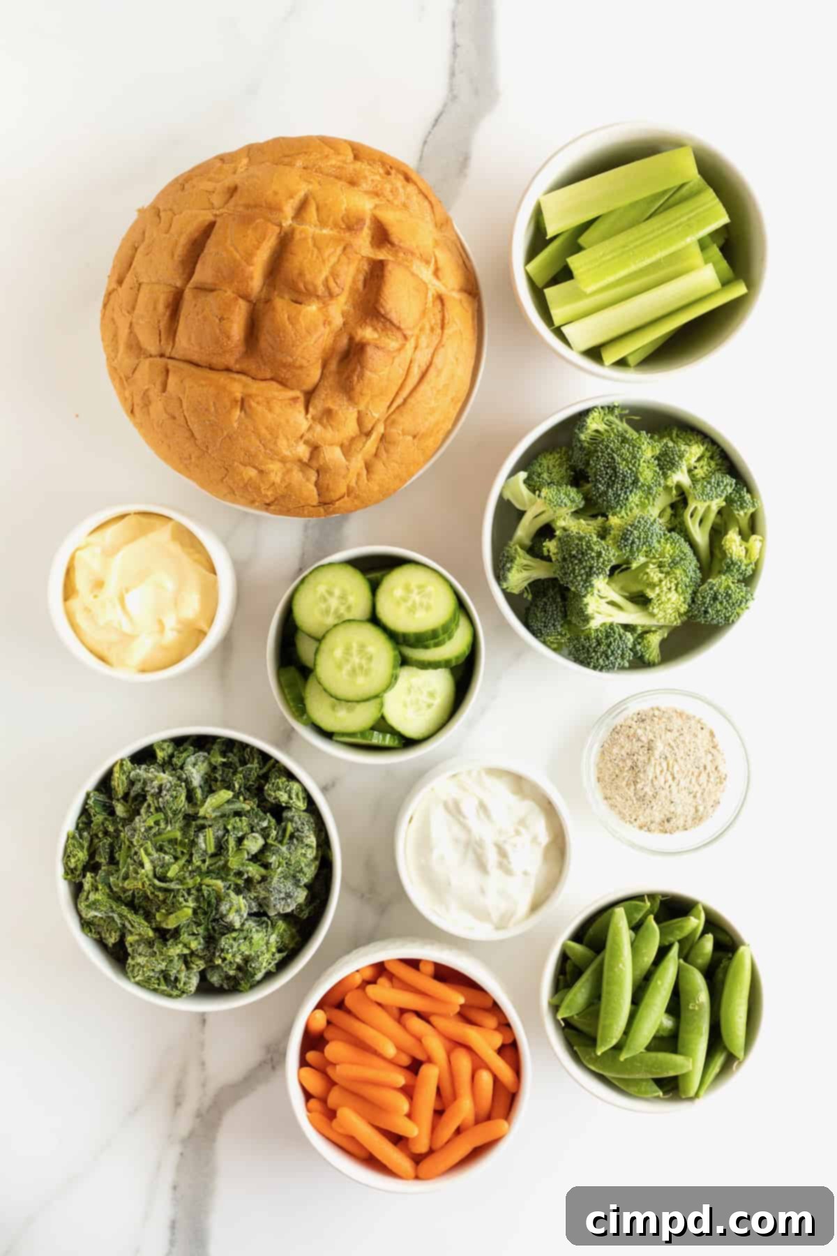 A selection of fresh ingredients including a bread bowl, cucumber slices, broccoli florets, carrot sticks, spinach, and cream cheese, laid out for a spinach dip preparation.