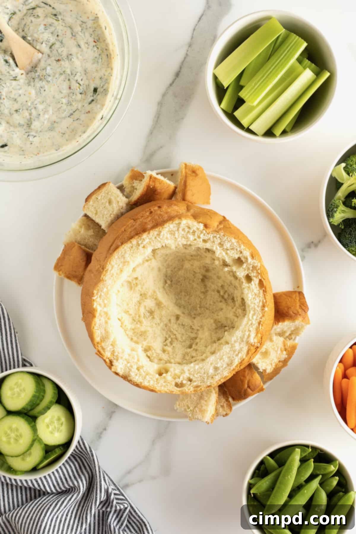 A hollowed-out bread bowl is presented on a white plate, surrounded by neatly cut bread chunks, ready for serving with dip.