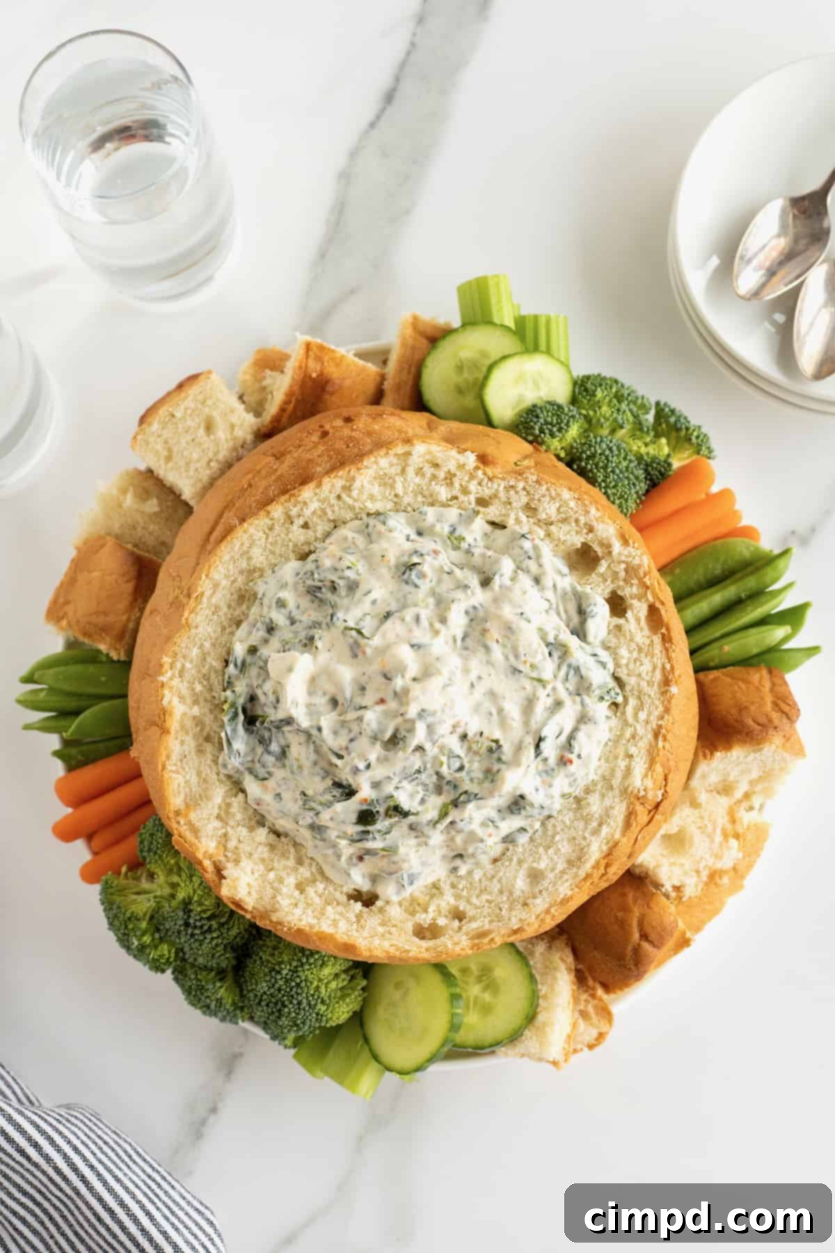 Creamy spinach dip nestled within a bread bowl, elegantly surrounded by an assortment of fresh vegetables and bread chunks, prepared for serving.