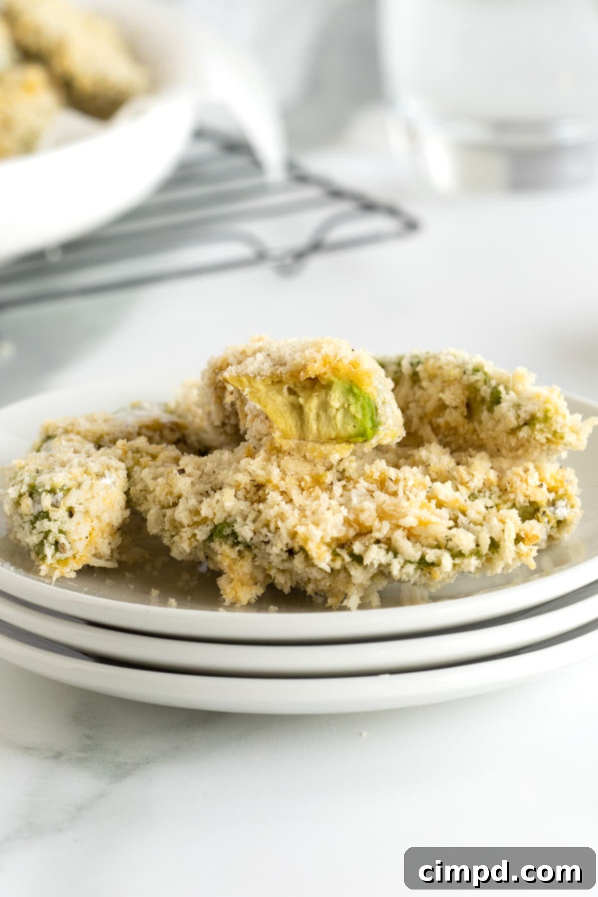 A cross-section of a baked avocado fry, showcasing its creamy interior and crisp breading, resting on a stack of three white plates.