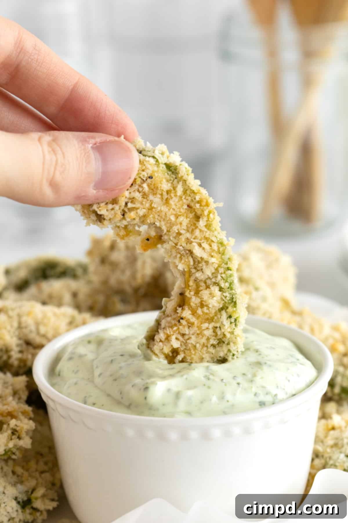 A baked avocado fry being dipped into a small bowl of fresh cilantro lime dipping sauce, highlighting its golden crust.