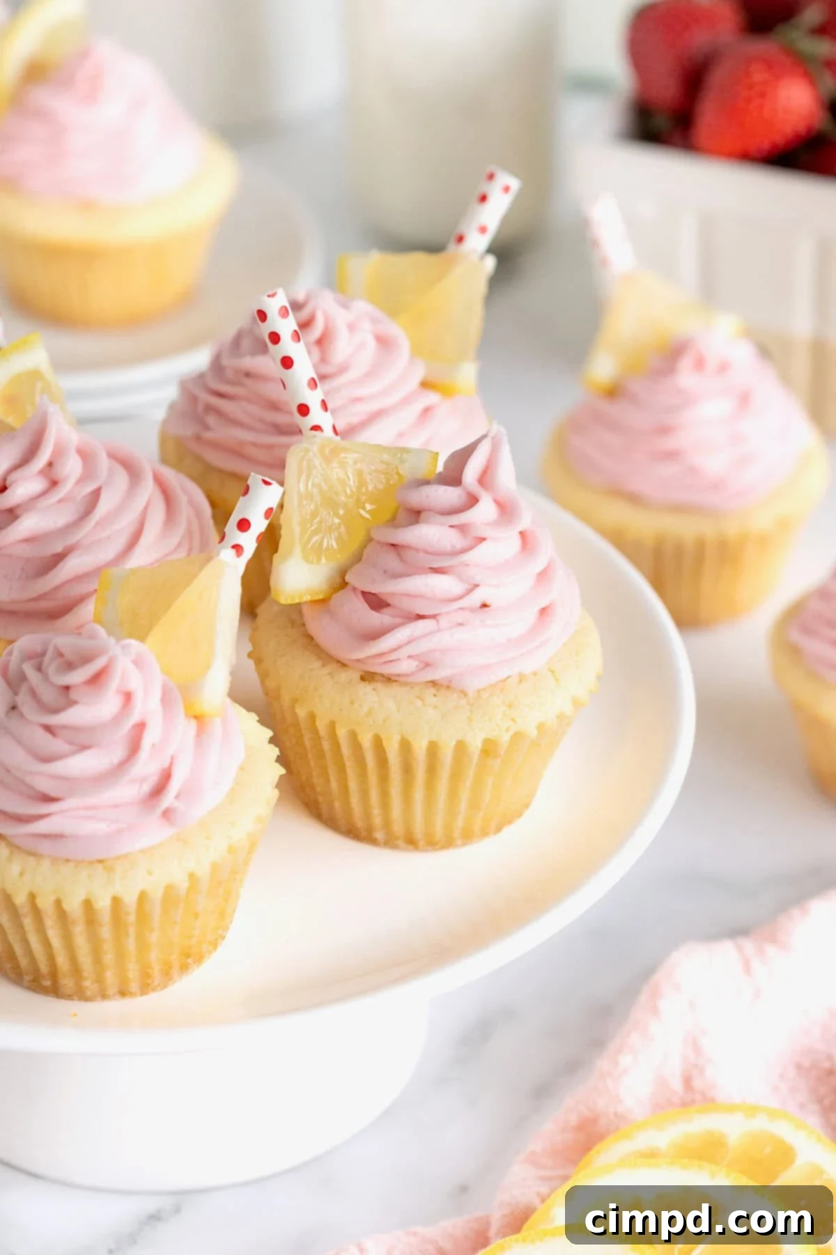 A white cake stand topped with beautifully frosted lemon cupcakes, each adorned with pink strawberry frosting, a small lemon triangle, and a charming red and white polka dot straw.