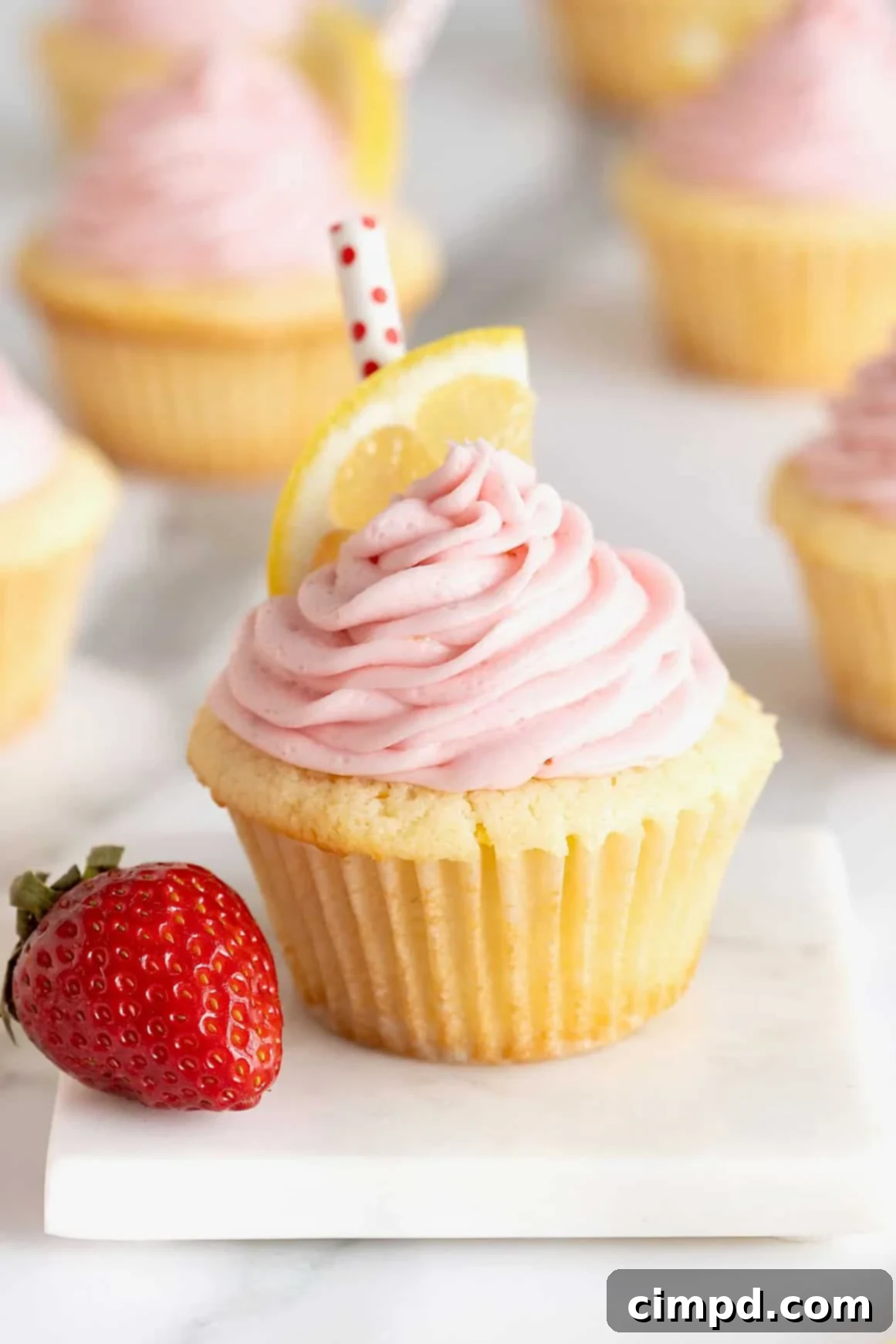 A beautifully presented strawberry lemonade cupcake resting on a white stone surface, with a whole fresh strawberry positioned to its left.