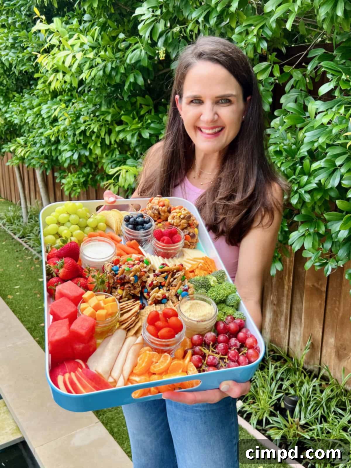 Picnic Perfection Platter 3 A smiling woman with long brown hair proudly holding a white-rimmed snack tray, artfully filled with a colorful medley of fresh fruits, crisp vegetables, and tempting no-bake delights, ready for an outdoor feast.