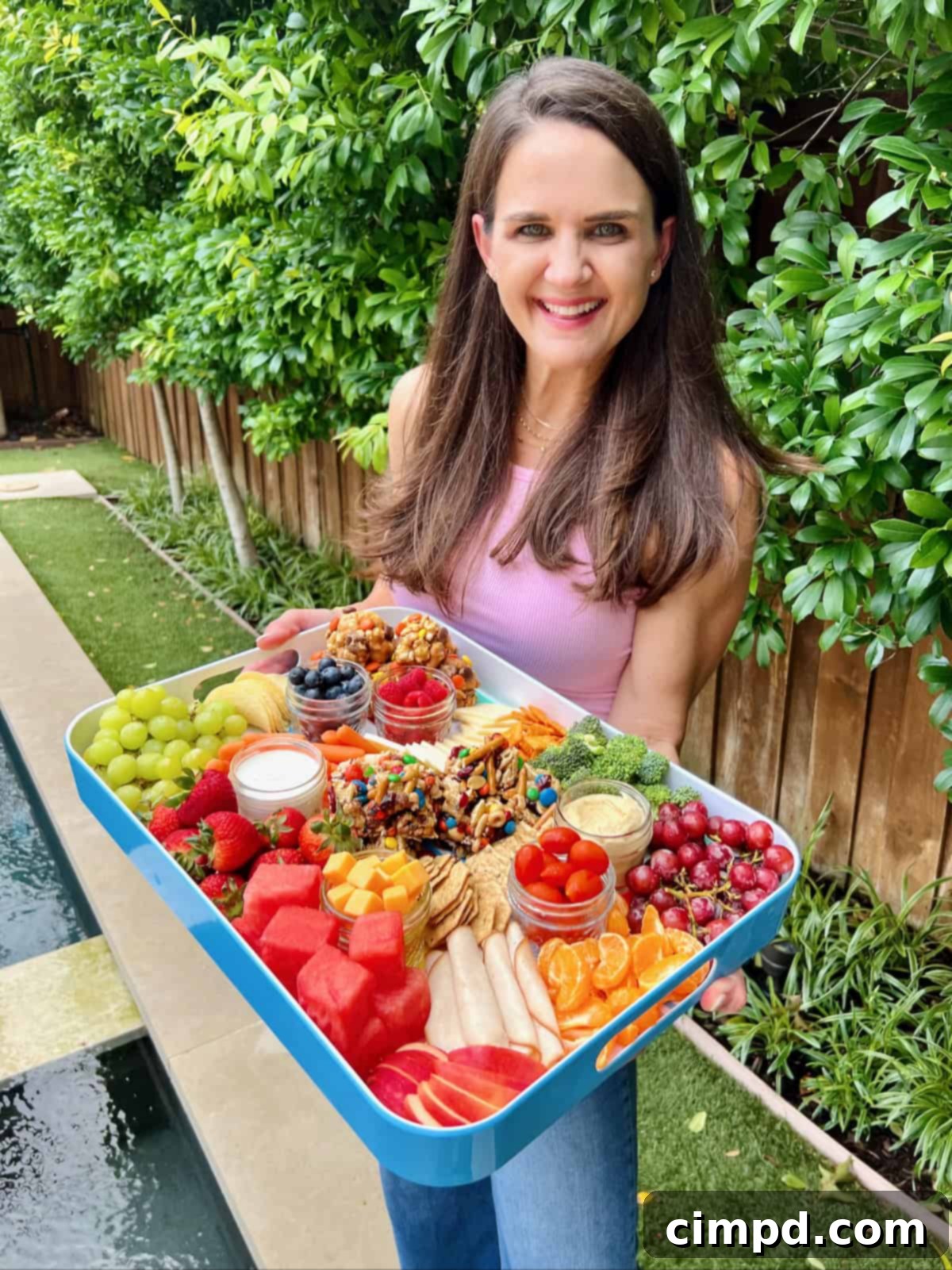 Picnic Perfection Platter 5 A beautiful woman with long brown hair gracefully presenting a white-rimmed snack tray, generously laden with an appealing assortment of fresh fruits, vibrant vegetables, and delicious no-bake snacks, showcasing a perfect balance of healthy and indulgent treats.