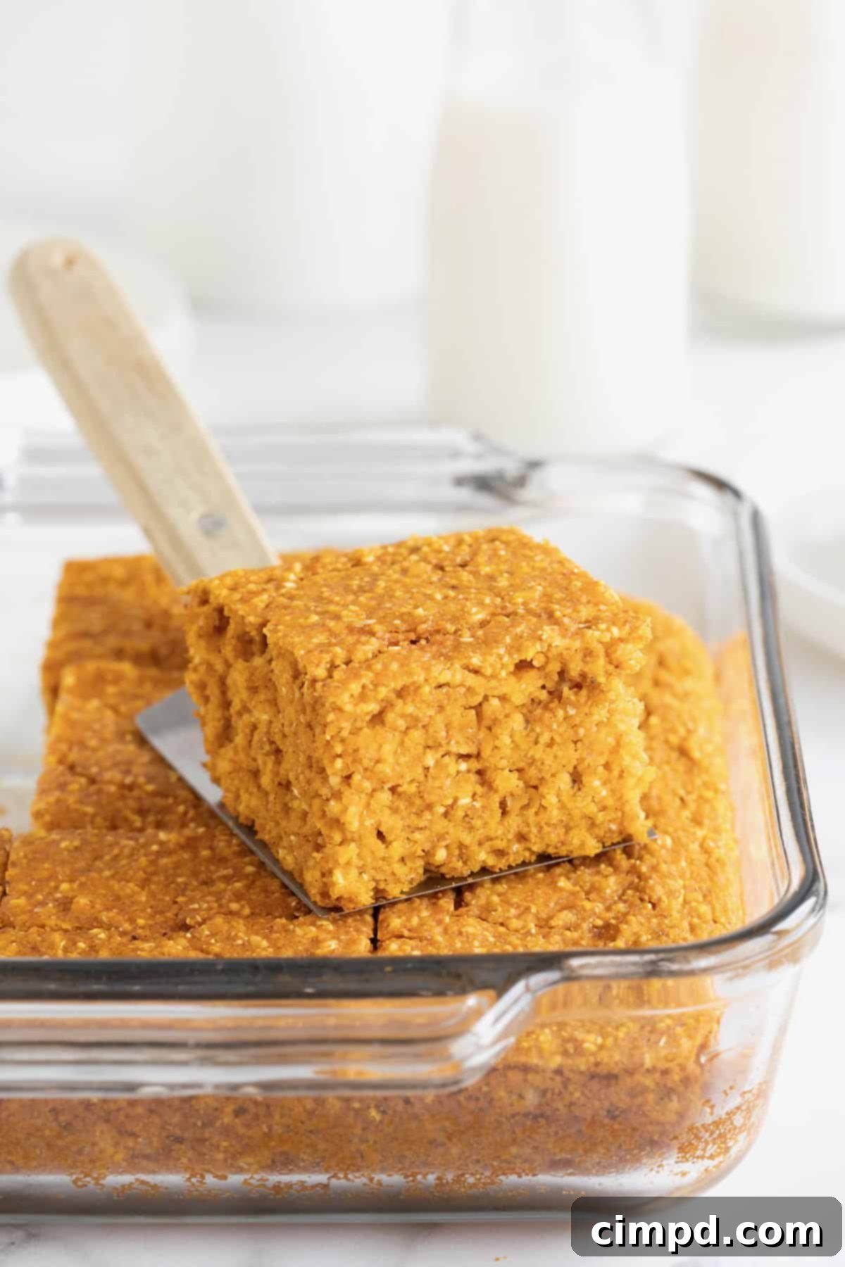 A beautiful glass baking dish filled with golden-brown whole wheat pumpkin cornbread, freshly baked and steaming. A metal spatula with a wooden handle is gently inserted, suggesting a slice is ready to be served.
