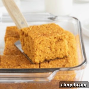 A glass baking dish of golden-brown whole wheat pumpkin cornbread. There is a metal spatula with a wood handle sticking out of the dish.