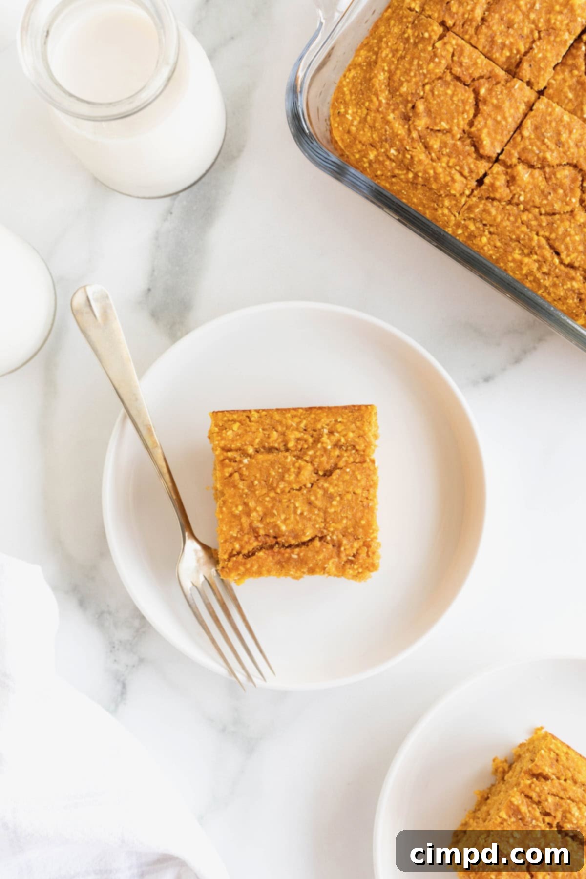 A freshly baked slice of pumpkin cornbread elegantly positioned on a small white plate, with a glass baking dish containing more cornbread visible in the soft background on a white marble counter.