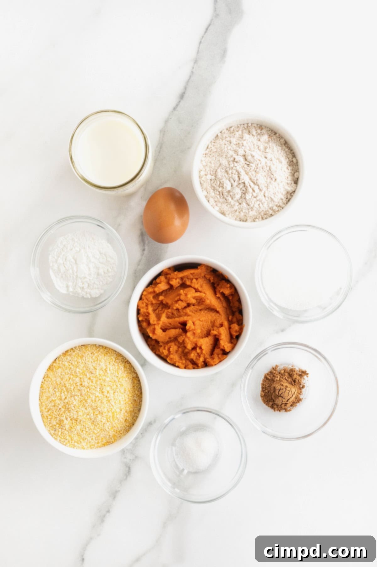 A curated selection of fresh ingredients laid out on a kitchen counter, ready for making whole wheat pumpkin cornbread. Ingredients include cornmeal, whole wheat flour, canned pumpkin puree, milk, an egg, and a jar of pumpkin pie spice.