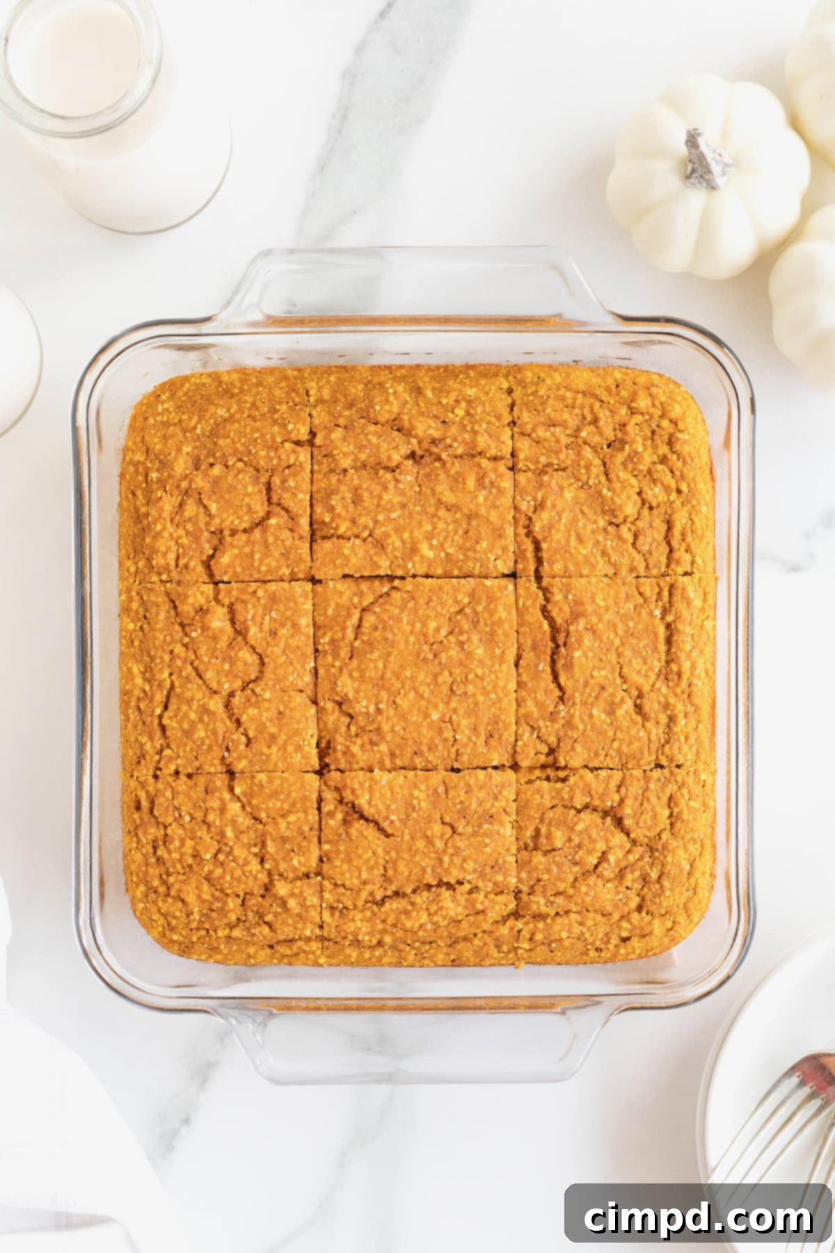 A freshly baked, golden-brown whole wheat pumpkin cornbread, neatly cut into nine perfect squares and arranged within its square glass baking dish, resting on a white marble counter.