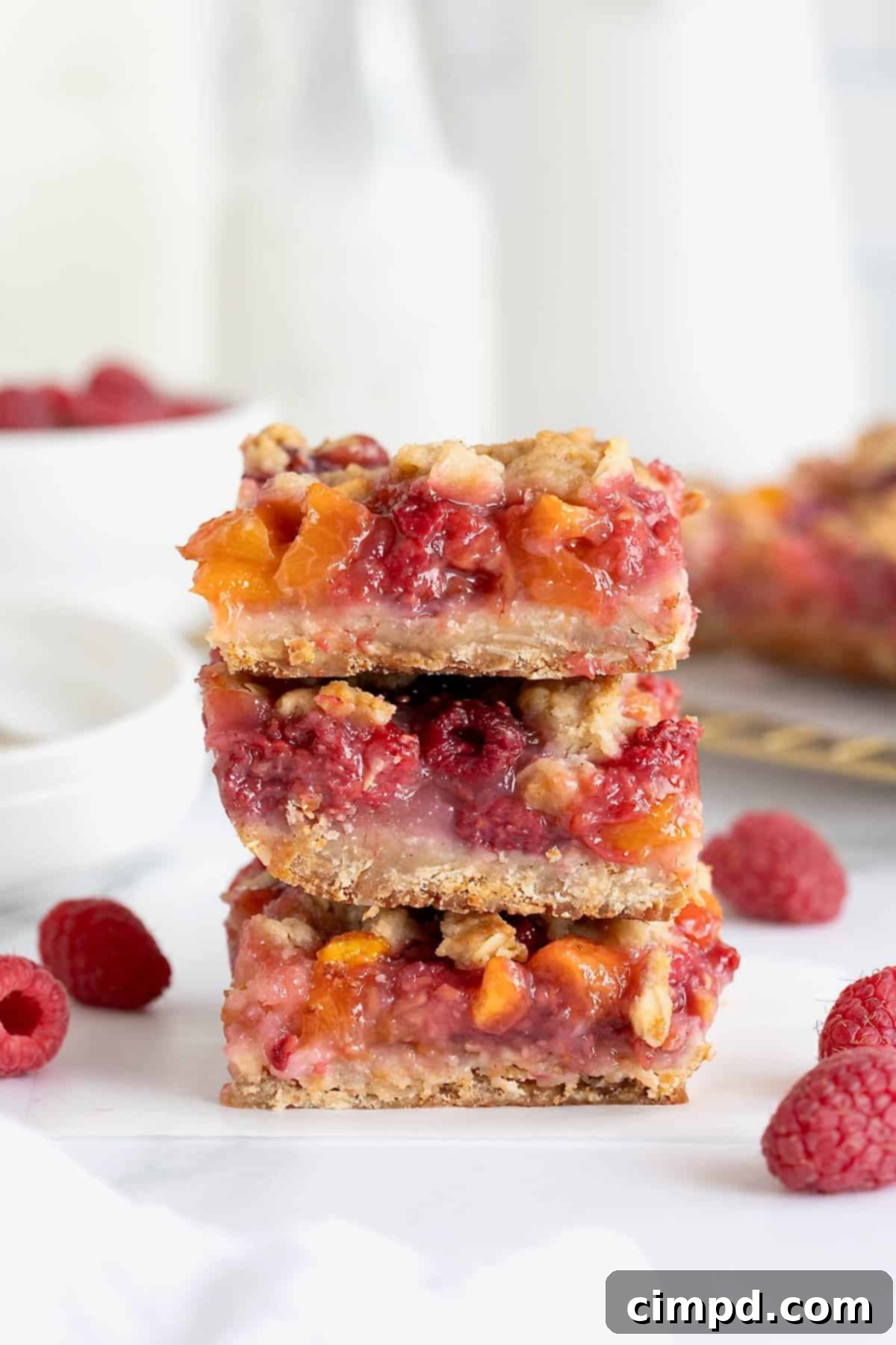 Rustic Peach Raspberry Crumbles 2 A stack of three raspberry peach crumb bars on a white marble counter. Raspberries are scattered around the bars.