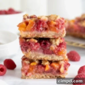 Golden Peach Raspberry Crumble Squares 13 A stack of three raspberry peach crumb bars on a white marble counter. Raspberries are scattered around the bars.