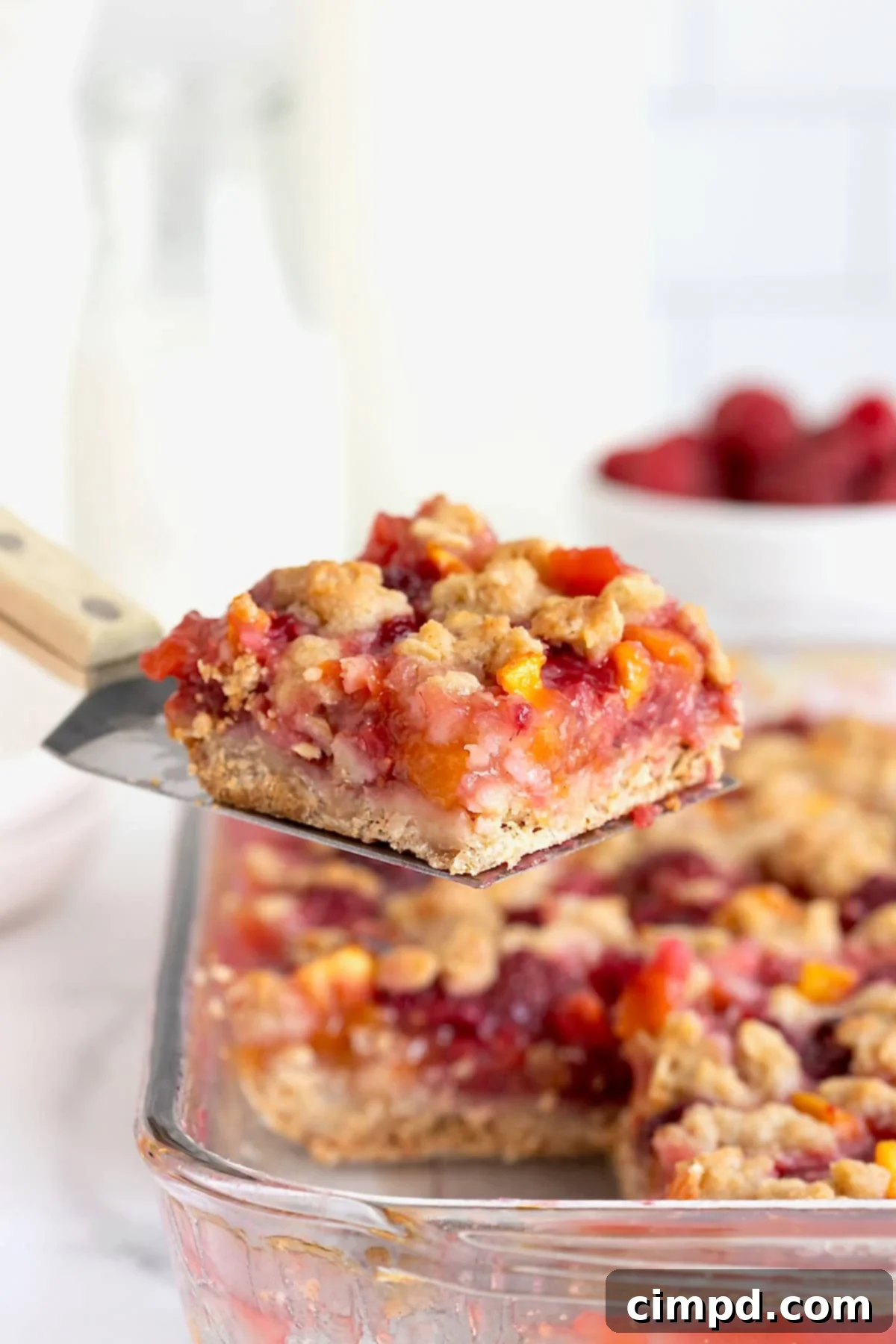 Golden Peach Raspberry Crumble Squares 3 A perfectly golden-brown peach raspberry crumb bar being carefully lifted from a baking dish with a spatula, showcasing its crumbly topping and juicy filling.