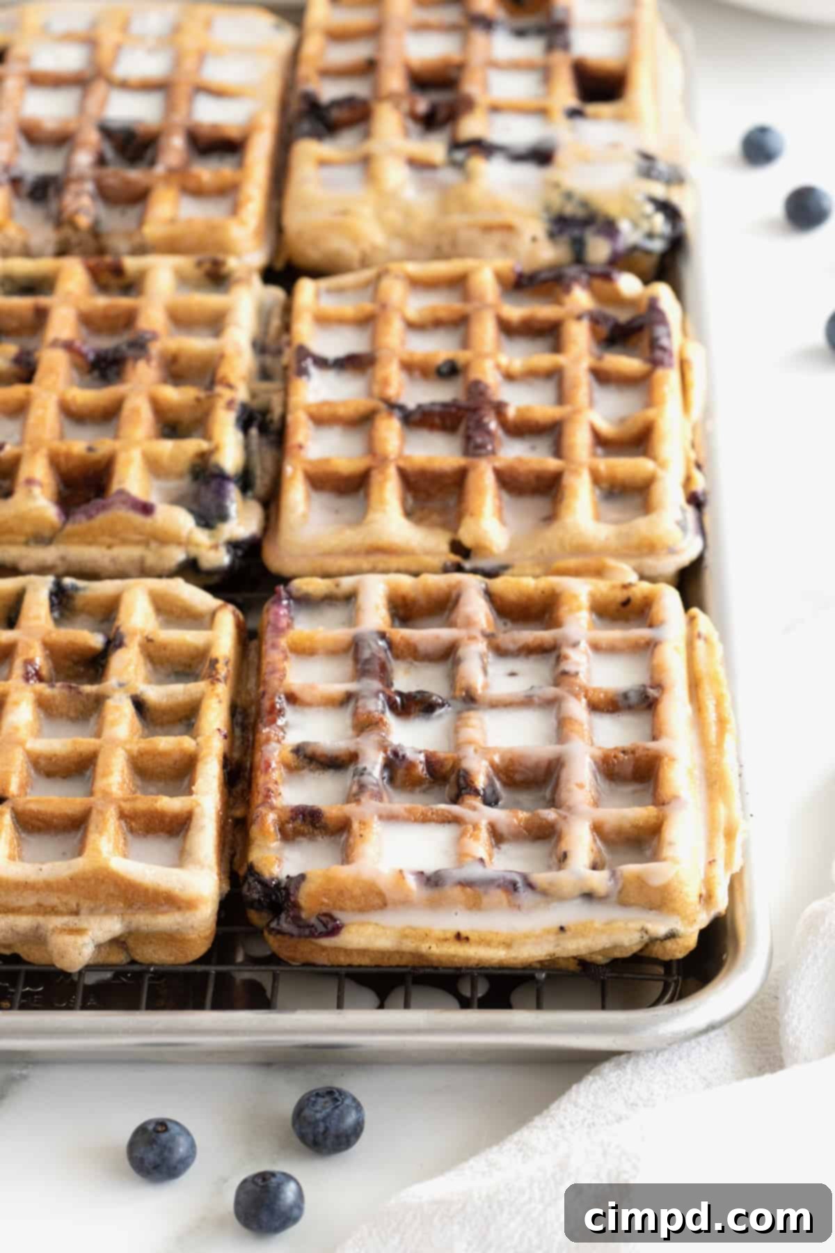 Six glazed blueberry waffles neatly arranged on an aluminum rimmed baking sheet, showcasing their golden-brown edges and luscious white glaze.