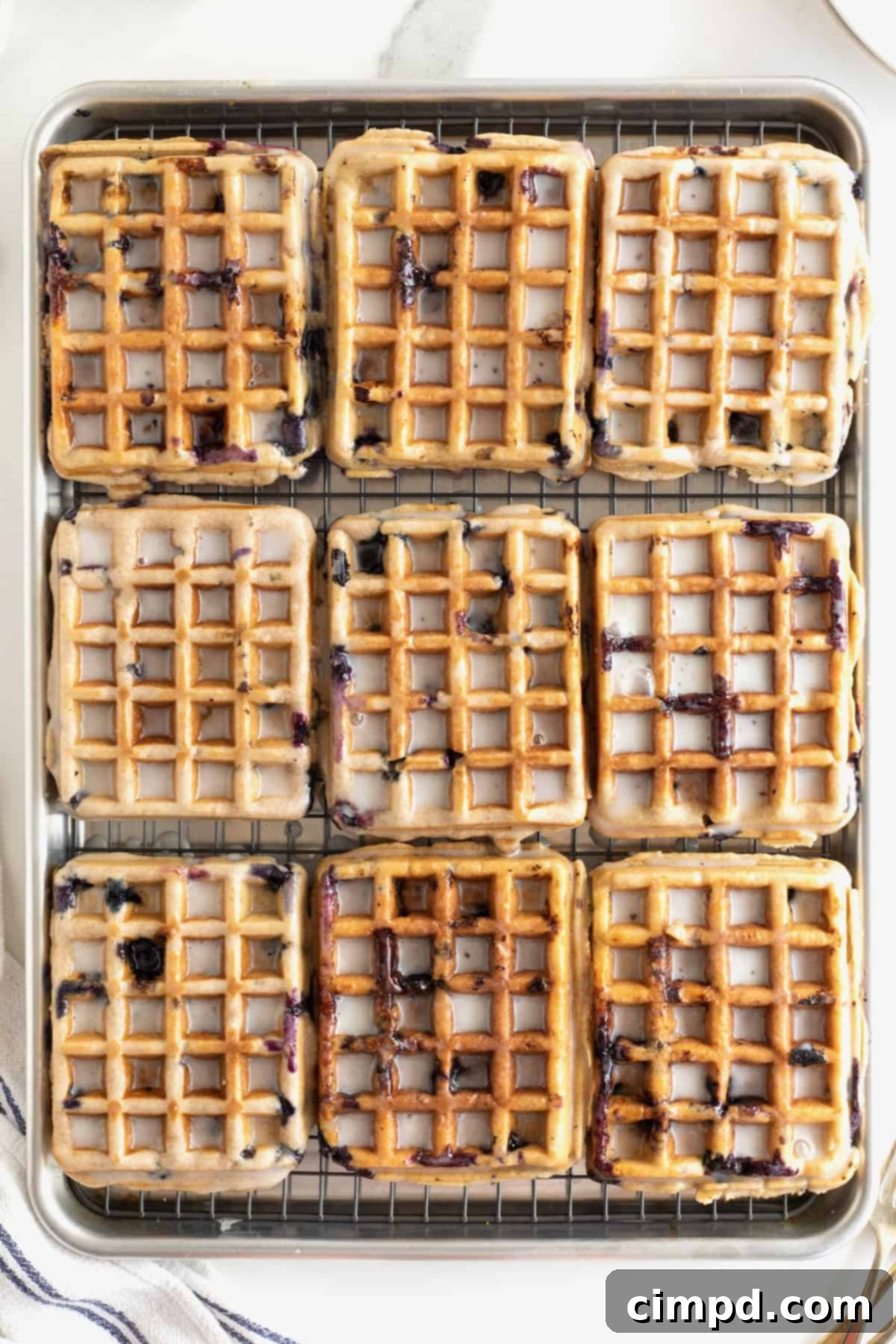 A full rack of nine blueberry waffles, glistening with a fresh white glaze, cooling over an aluminum rimmed baking sheet.
