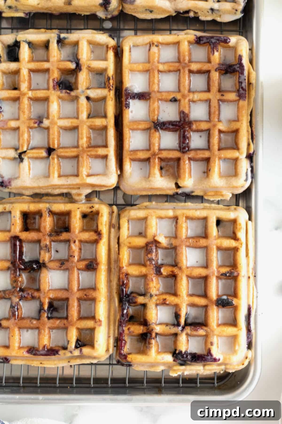 Six glazed blueberry waffles displayed on an aluminum rimmed baking sheet, ready for storage or serving, with a slight purplish tint from the blueberries peeking through the glaze.