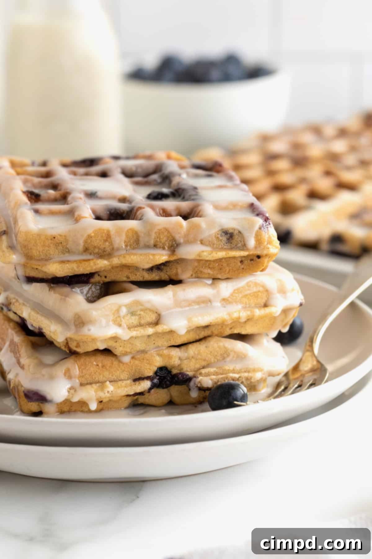 A stack of three perfectly glazed blueberry waffles rests on a small white plate, with a fork poised, inviting a taste.