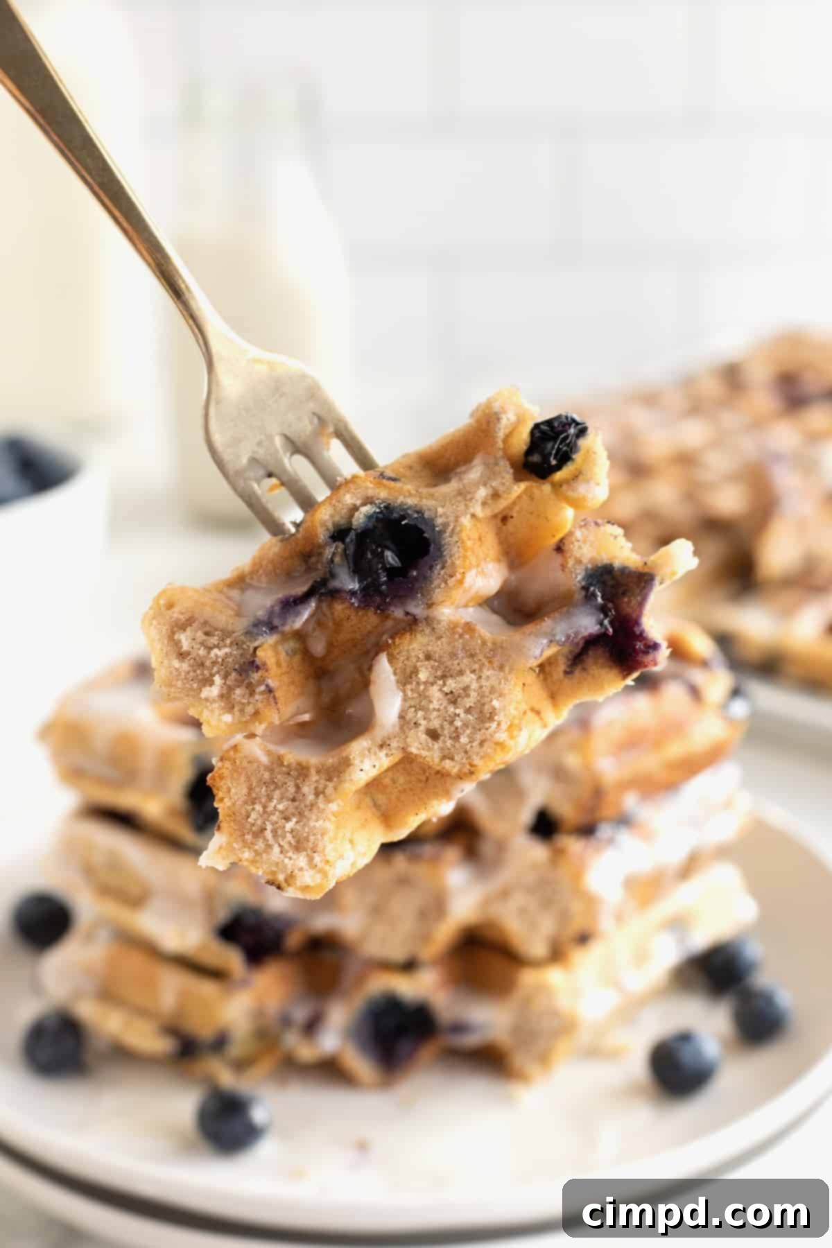 Three glazed blueberry waffles stacked on a white plate, with a fork holding a perfect bite in the foreground, highlighting the fluffy texture and juicy blueberries.