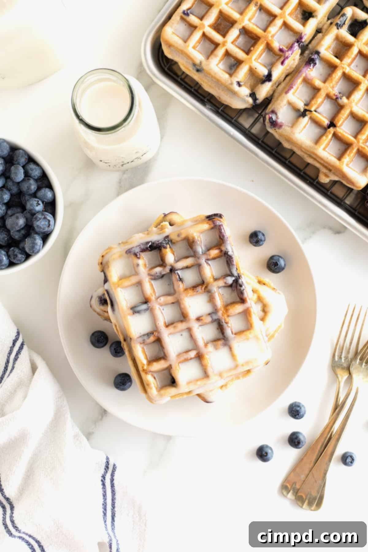 A single beautifully glazed blueberry waffle rests on a white plate, surrounded by scattered fresh blueberries and two elegant gold-toned forks.