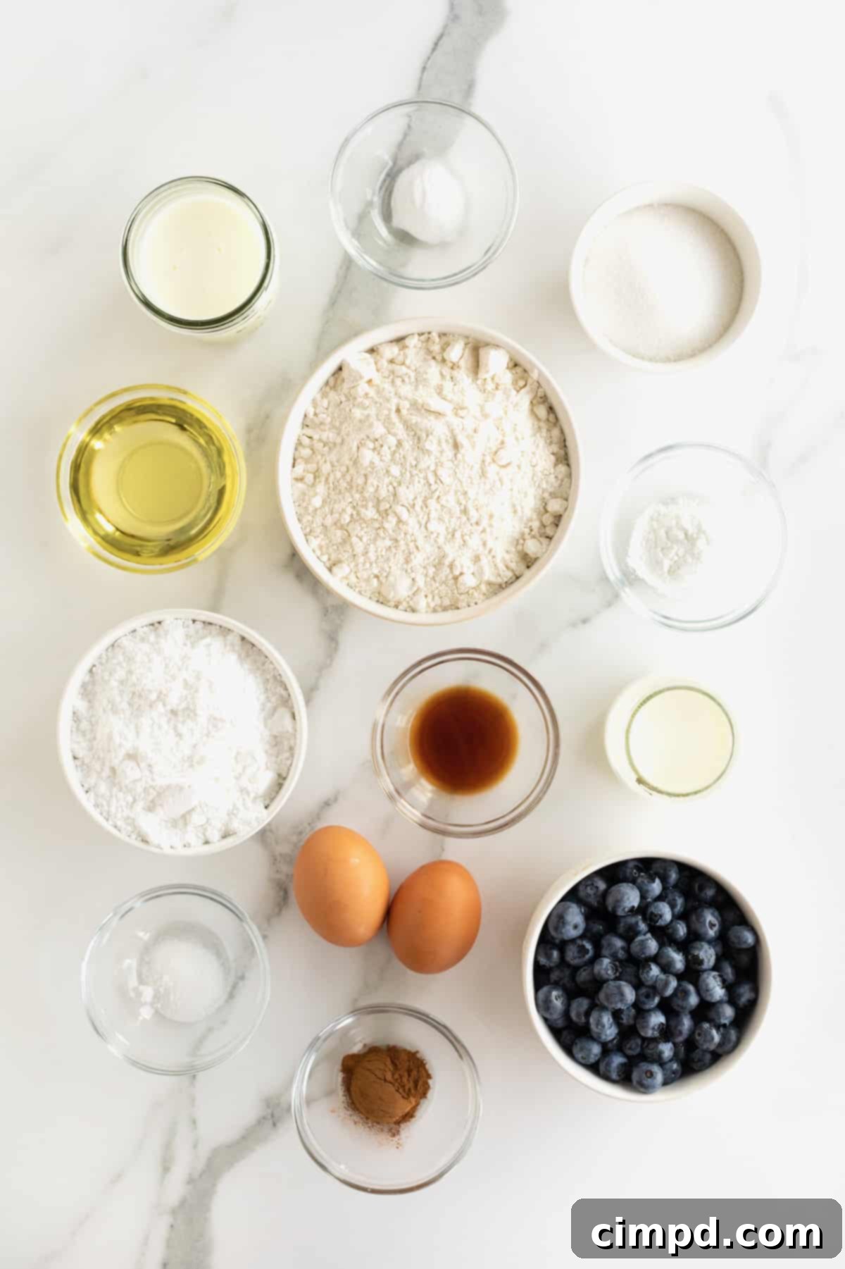 A beautiful display of all the essential ingredients for blueberry cake waffles, meticulously arranged in small glass dishes on a pristine white marble counter, ready for preparation.