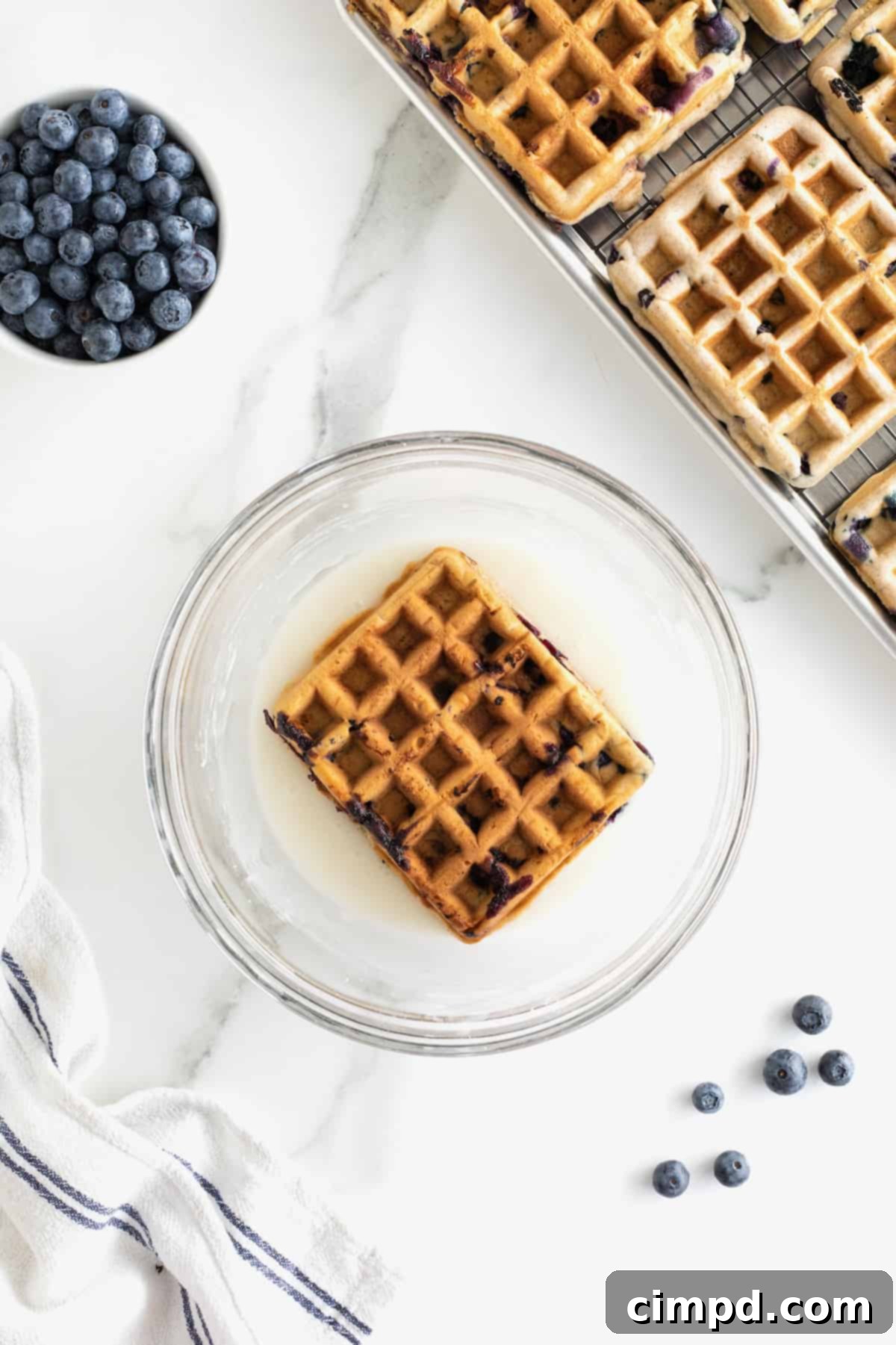 A golden blueberry waffle being gently dipped into a glass bowl of luscious white glaze, ensuring a rich coating.