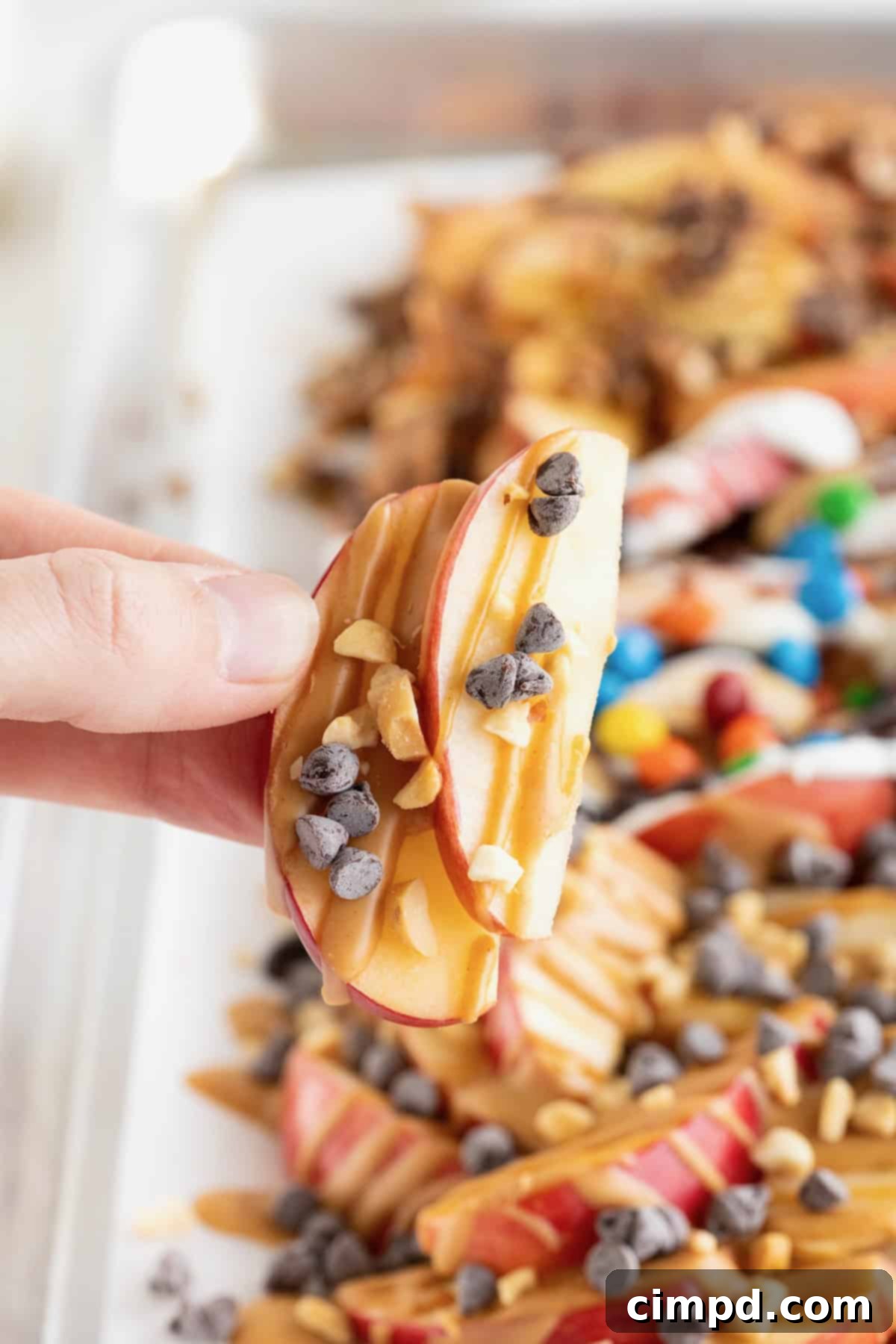 Loaded Apple Slices 3 A hand holding two thin slices of apple coated in caramel and topped with mini chocolate chips between the thumb and forefinger.