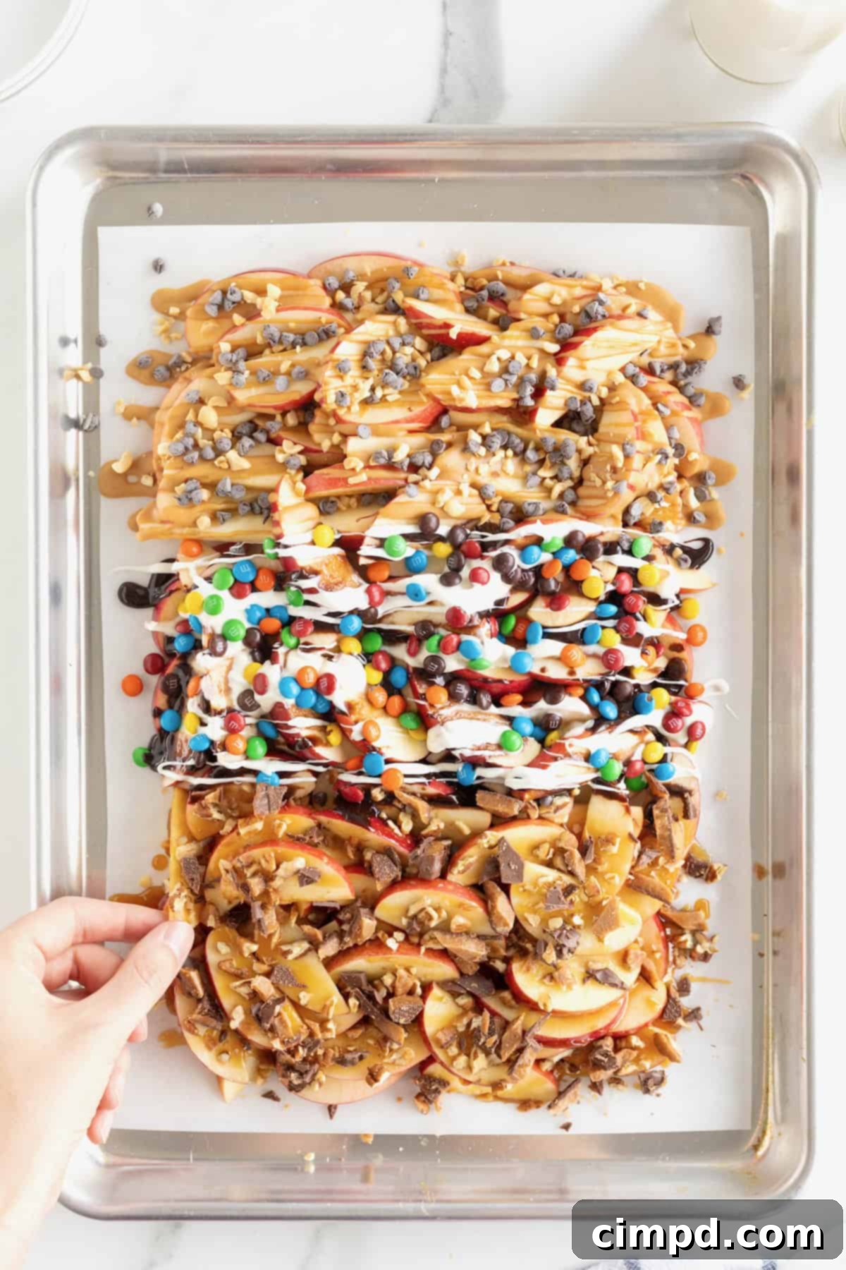Loaded Apple Slices 9 A hand reaching in to take a piece of apple nachos on a parchment lined aluminum baking sheet.