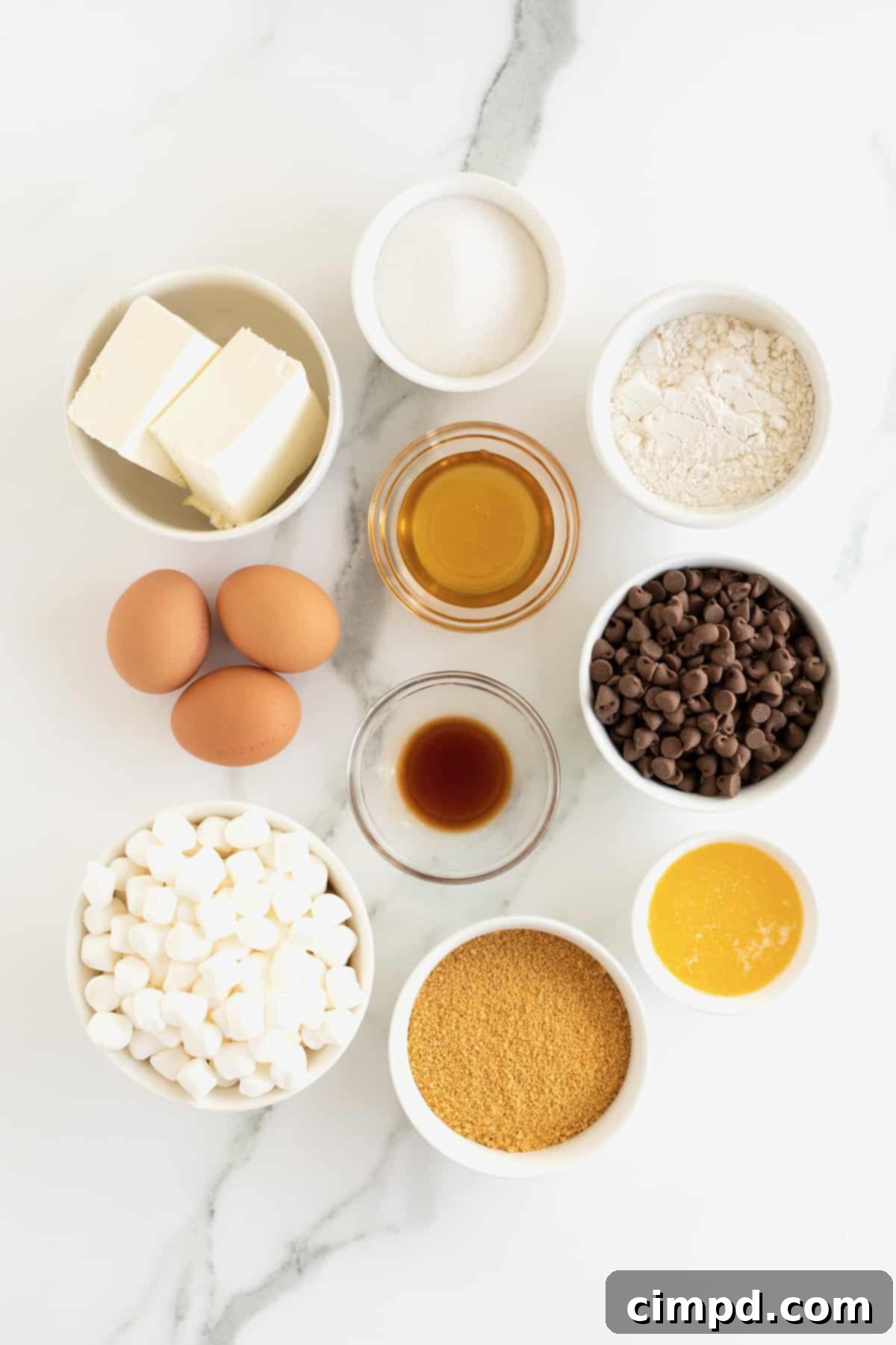 Marshmallow bits, chocolate chips, butter eggs, cream cheese, flour and sugar tin small glass dishes on a white marble counter.