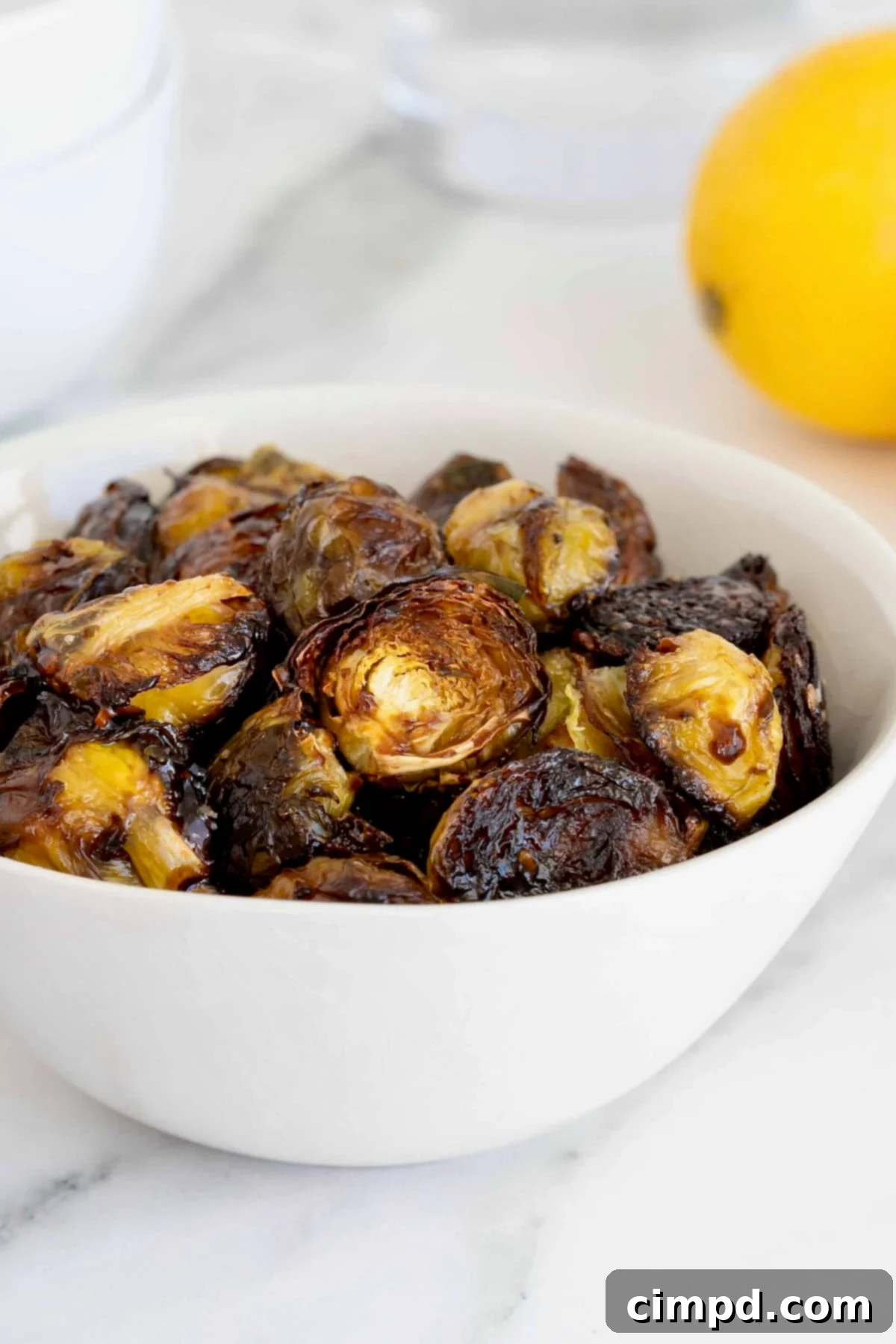 A bowl of charred saucy Brussels sprouts in a white serving bowl on a white marble counter. A lemon sits to the upper right corner in the background, suggesting fresh ingredients.