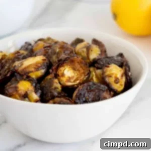 A bowl of charred saucy Brussels sprouts in a white serving bowl on a white marble counter. A lemon sits to the upper right corner in the background.