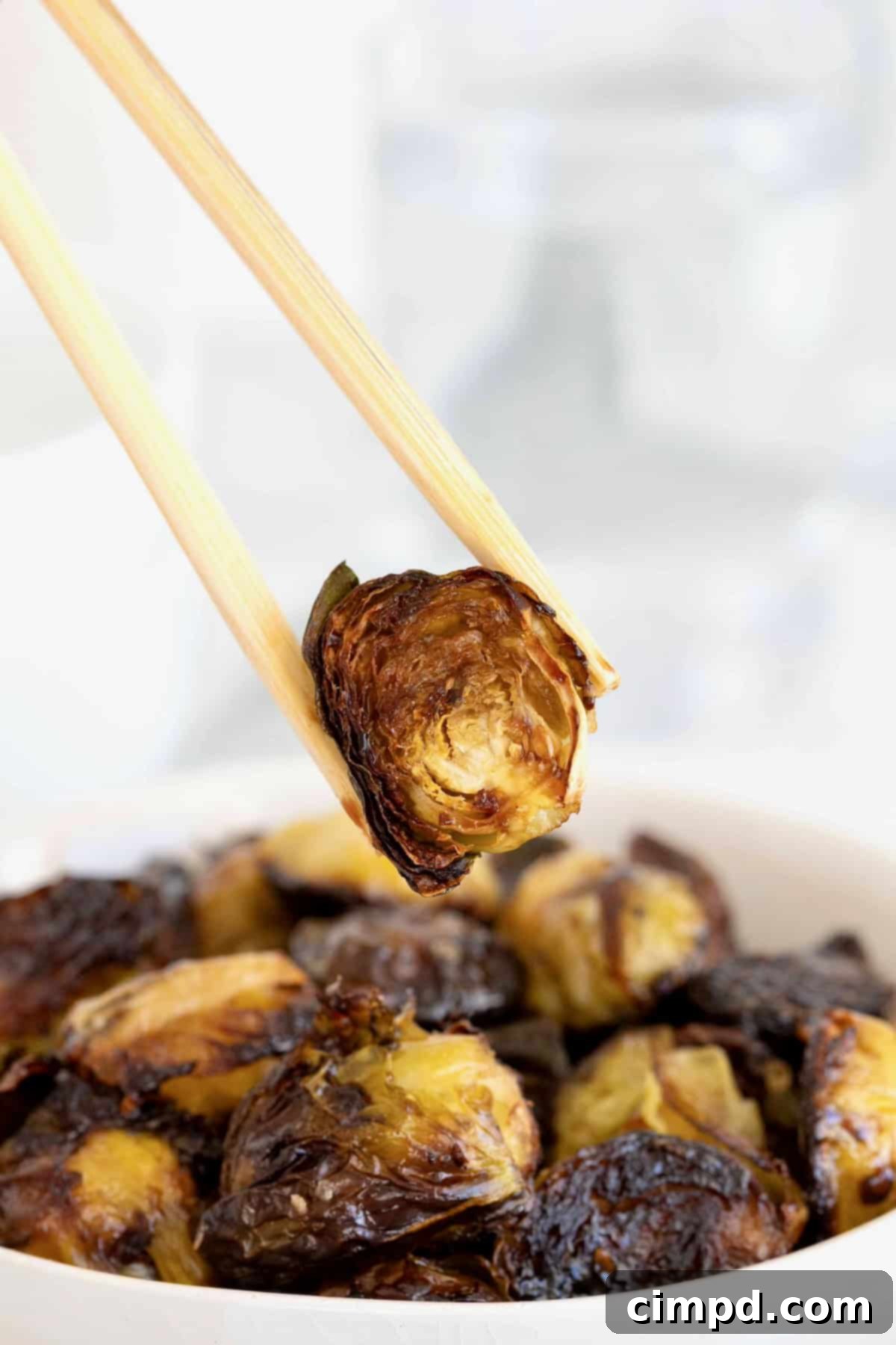 A pair of wooden chopsticks holding a slightly charred Brussels sprout half with sauce, highlighting its crispy texture.