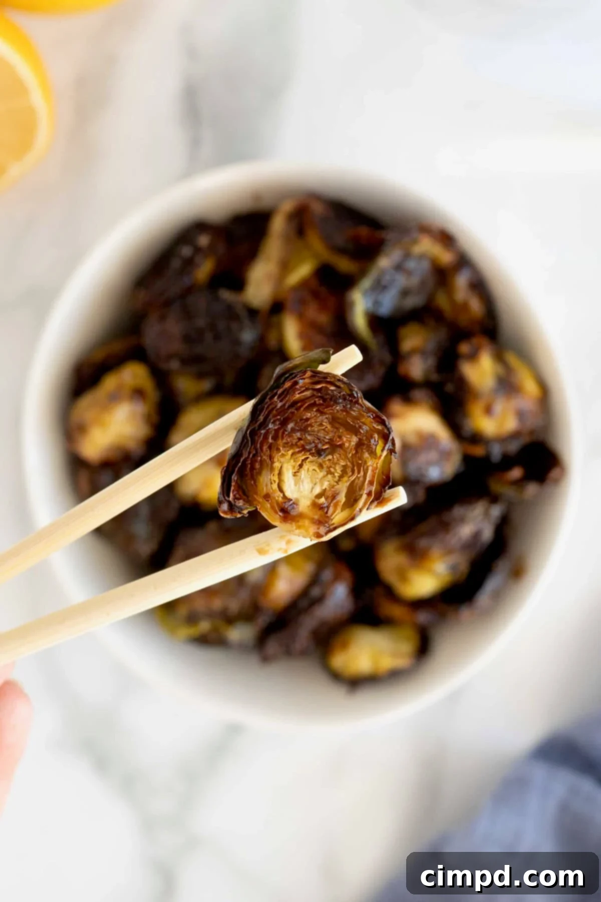A pair of wooden chopsticks holding a slightly charred Brussels sprout half with sauce, showing the glistening glaze.