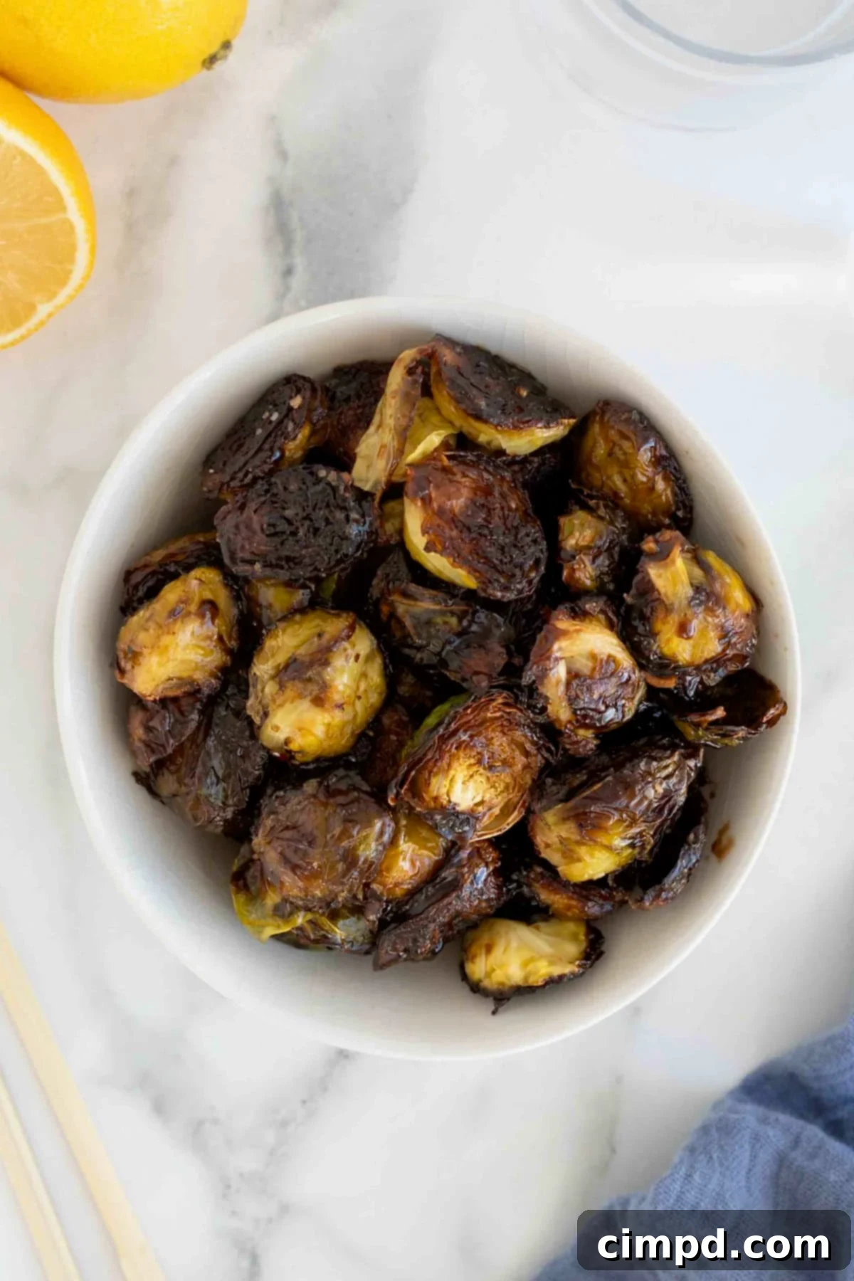 A bowl of charred saucy Brussels sprouts in a white serving bowl on a white marble counter. Half of a lemon sits to the upper left corner in the background, adding a fresh touch.